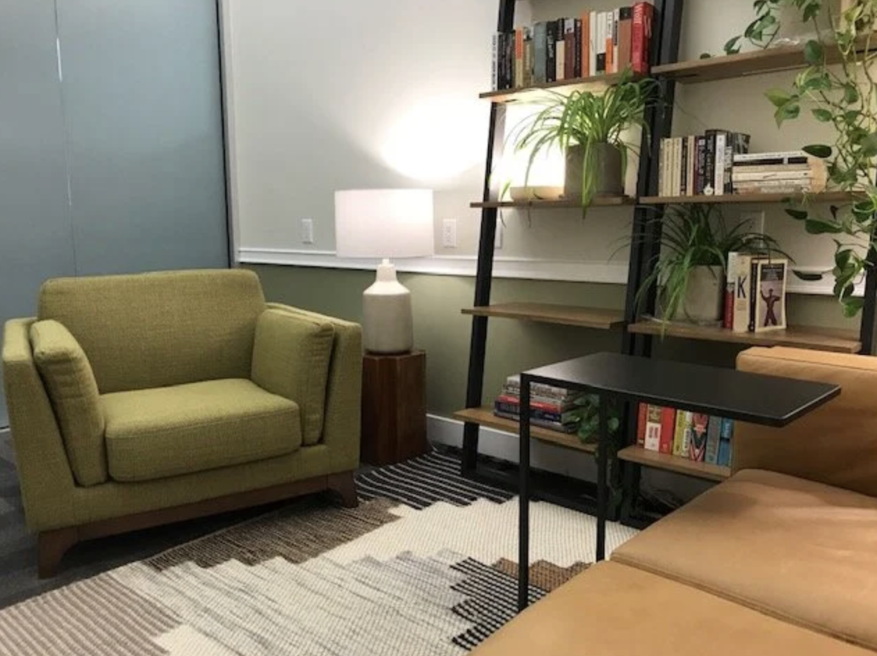 Living room with a green armchair, wooden side table with white lamp, black bookshelf filled with books and plants, beige modern sofa, striped rug, and a white wall with a sliding door.