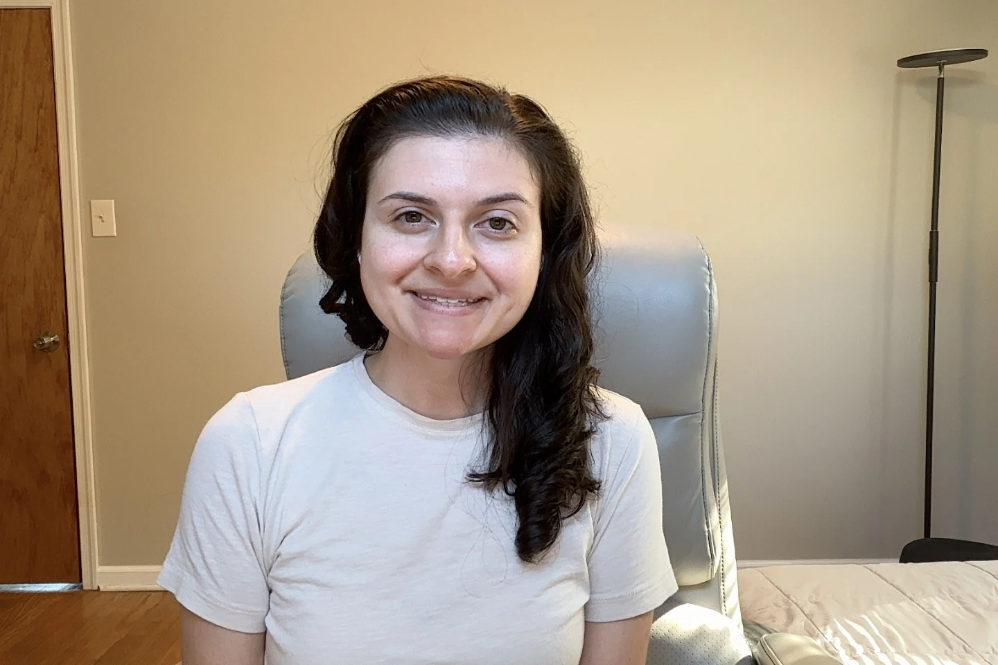 A woman with shoulder-length dark hair wearing a white t-shirt, sitting in a beige leather chair in a room with neutral-colored walls, a wooden door, a floor lamp, and a bed with a beige blanket.