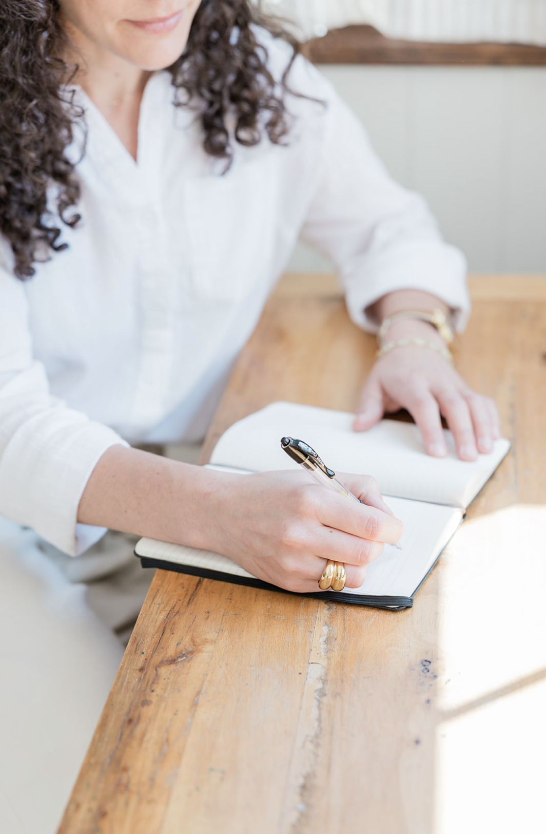 A woman with curly brown hair writing in a notebook with a black pen on a wooden table, wearing a white long-sleeve shirt and gold jewelry.