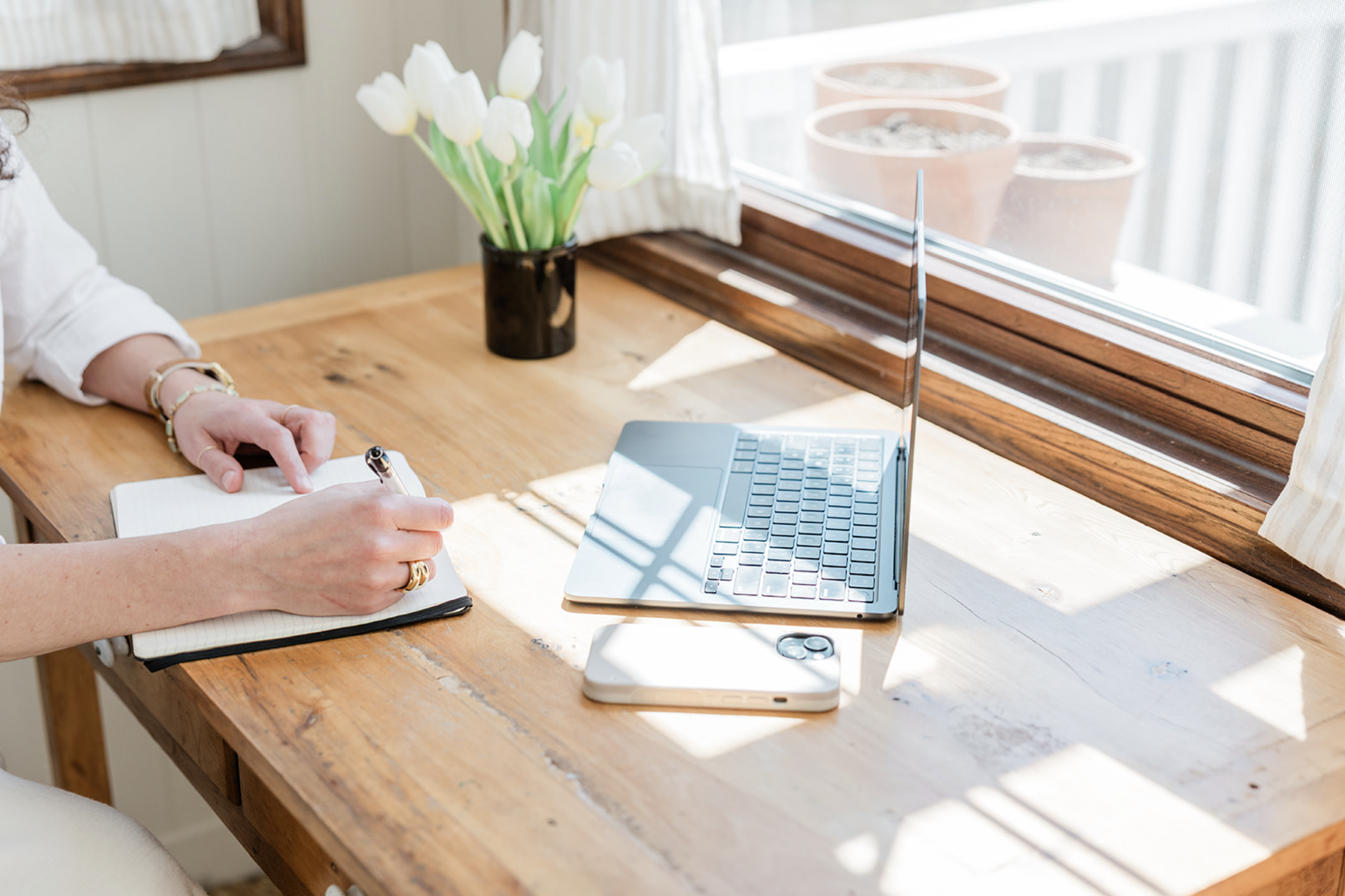 A person sitting at a wooden table, writing in a notebook with a pen, near a laptop, with a vase of white tulips and a window with curtains in the background.