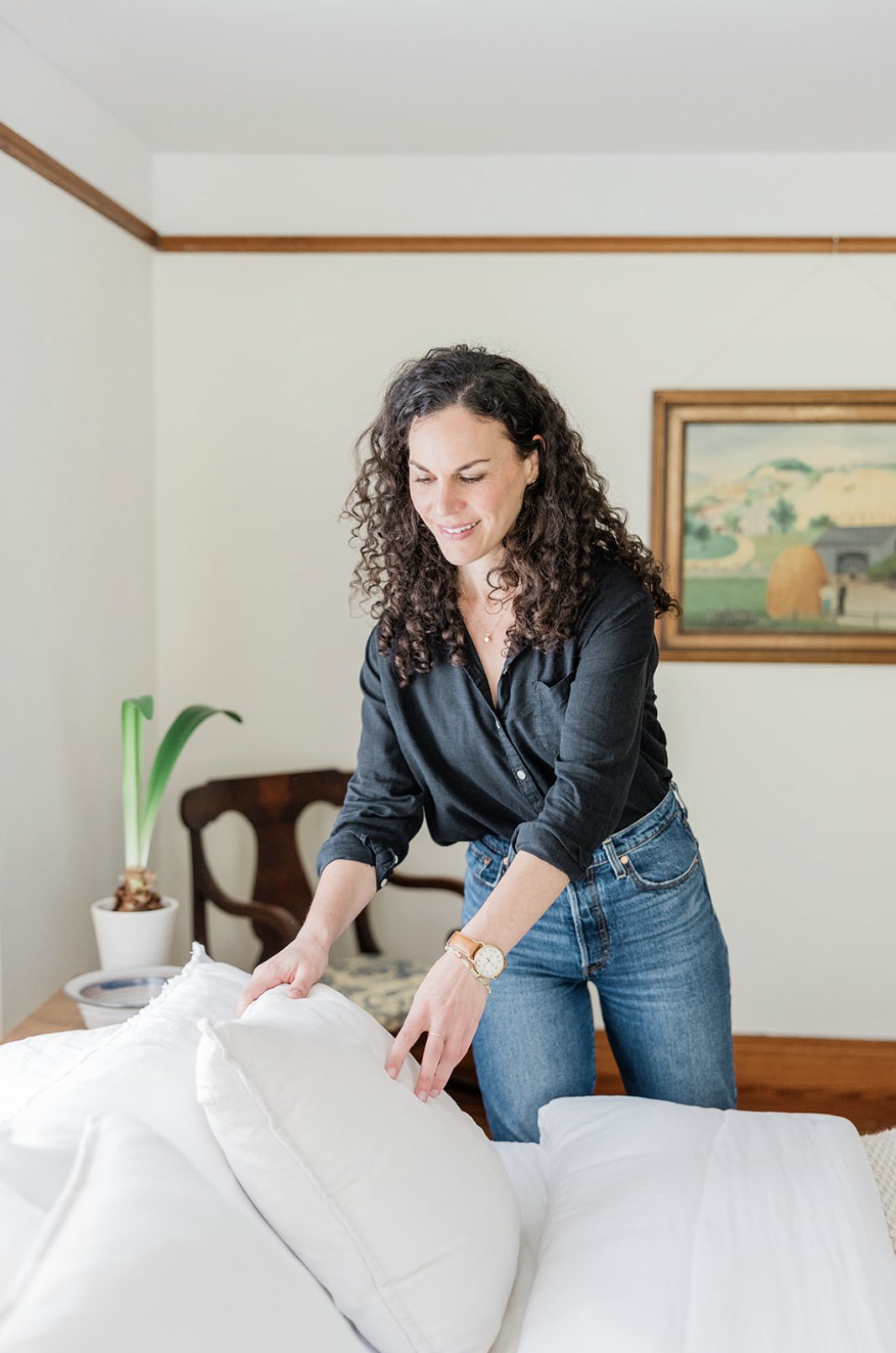 A woman with curly brown hair, wearing a black shirt and blue jeans, making the bed with white bedding in a bright, stylish bedroom.