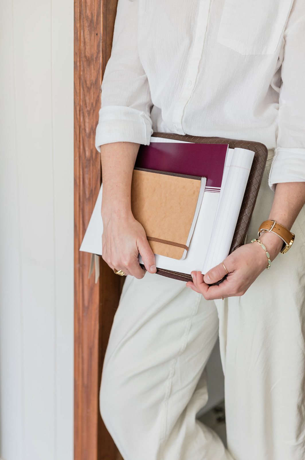 Person holding a brown leather folder and notebooks, dressed in a white shirt and beige pants, standing next to a wooden wall.