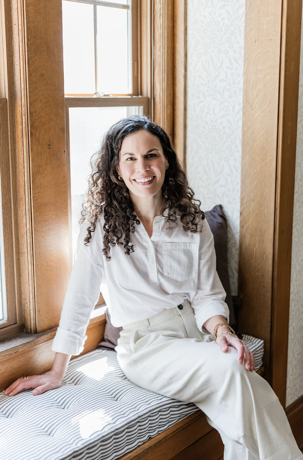 A woman with curly dark hair smiling while sitting on a window seat in a room with wooden trim and light-colored wallpaper.