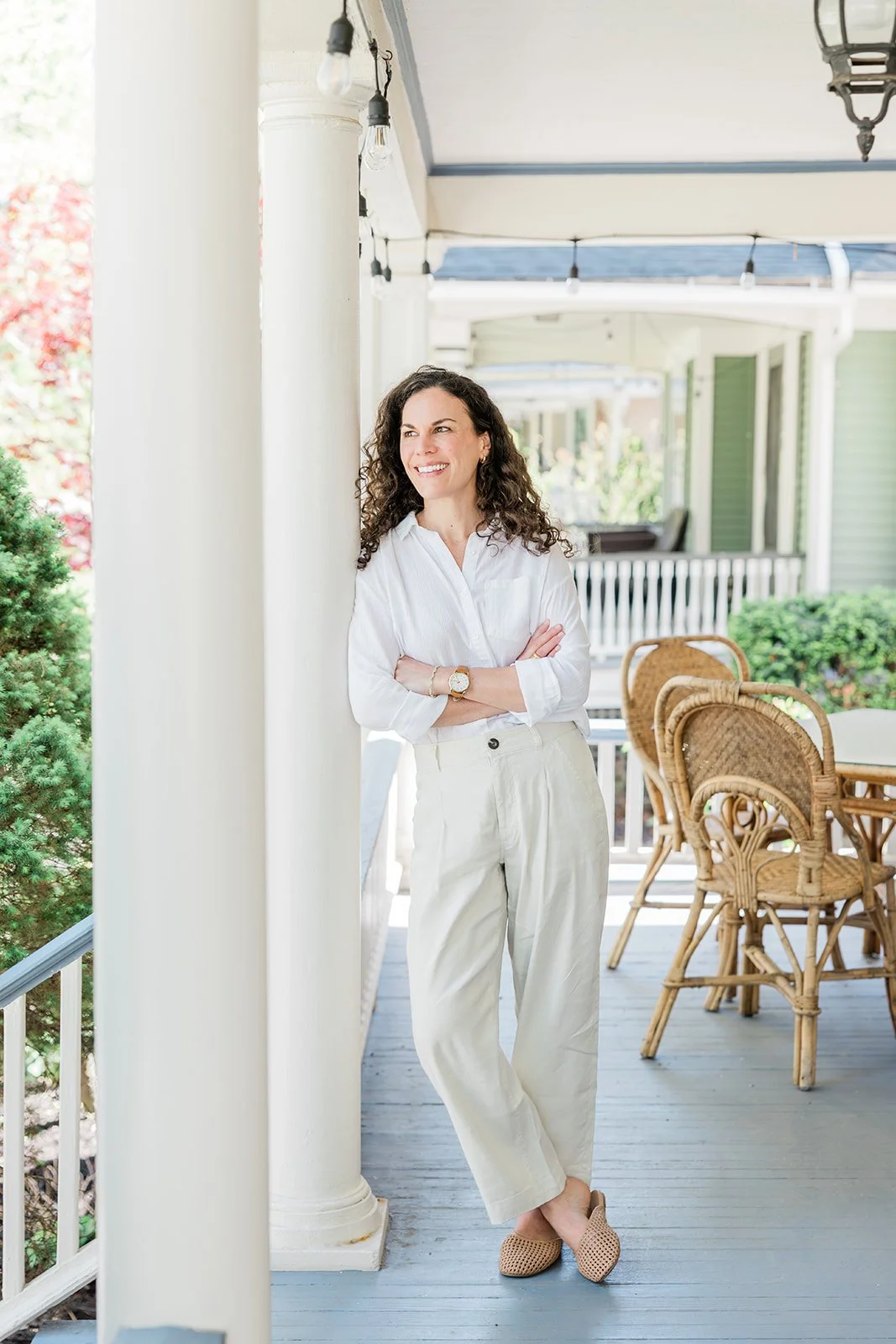 A woman with curly dark hair, wearing a white button-up shirt and cream-colored pants, stands on a porch with her arms crossed, smiling and looking to her left.