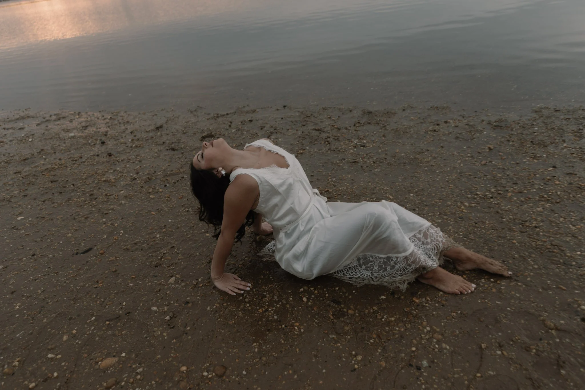 A woman in a white dress lying on a sandy beach near the water, with her head tilted back and eyes closed.