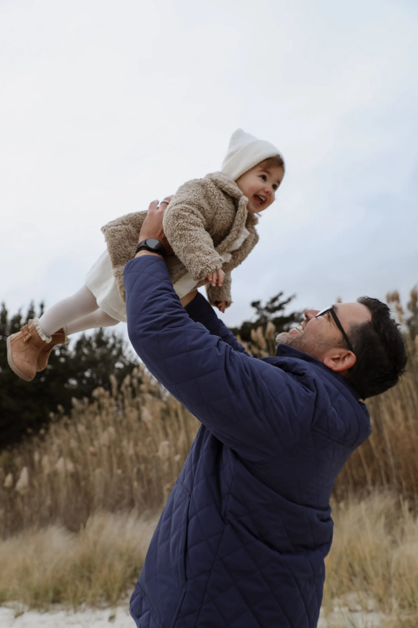 A man lifting a young girl in the air outdoors during winter, with trees and grasses in the background.