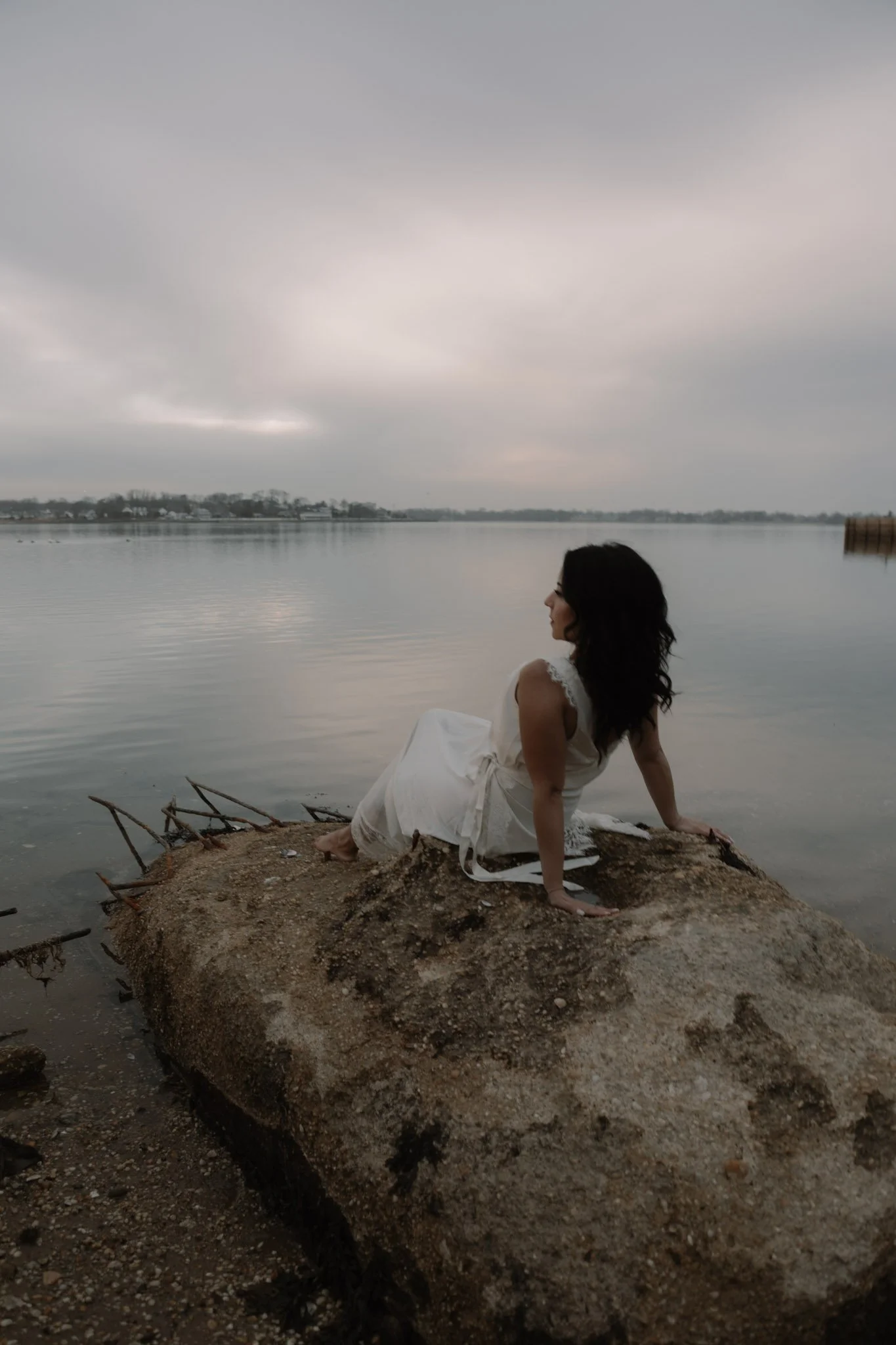 A woman in a white dress sitting on a large rock by a body of water, with an overcast sky overhead.