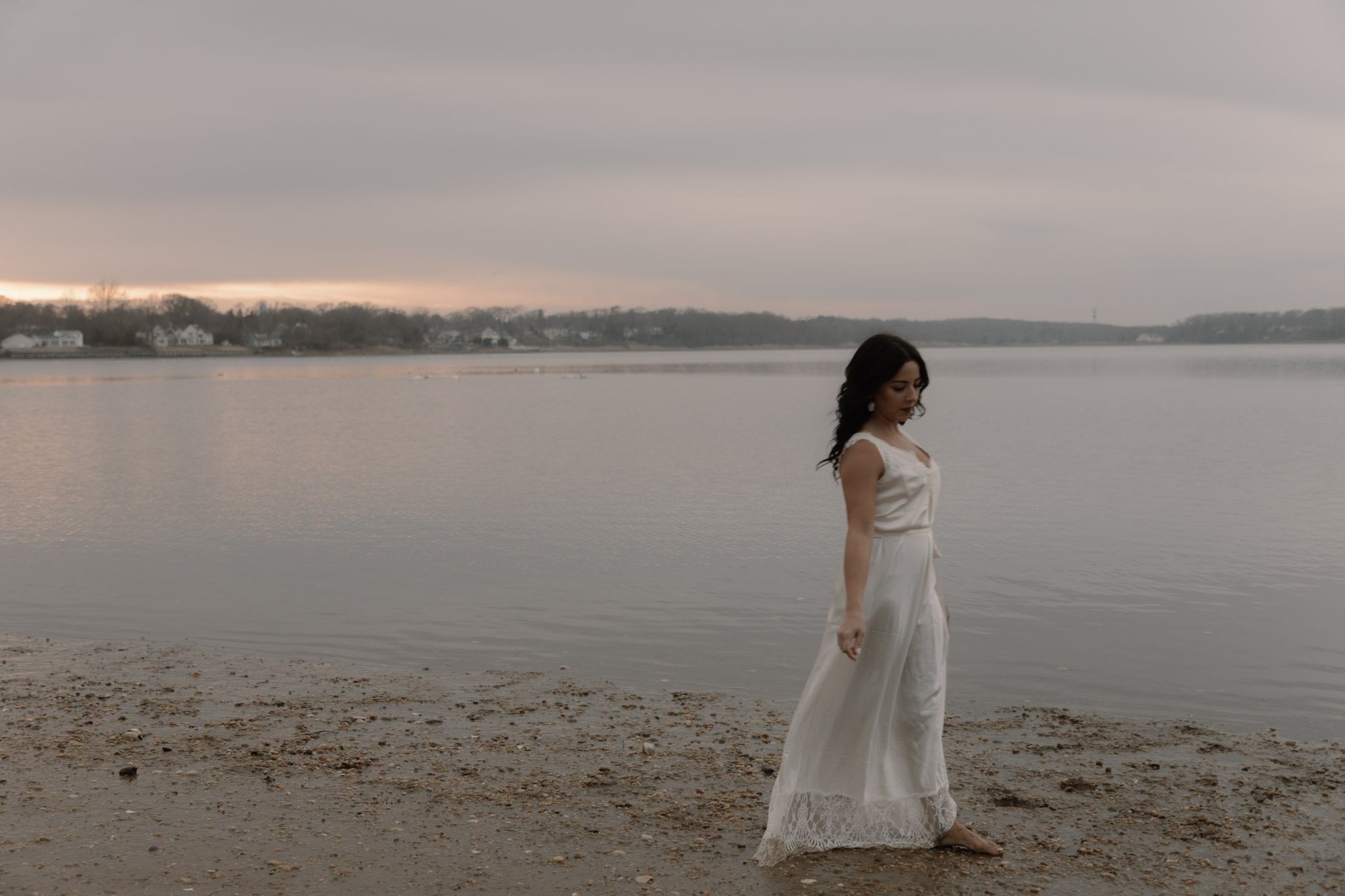 A woman in a white dress standing barefoot on a sandy beach near a body of water, with overcast skies and houses visible in the distance across the water.