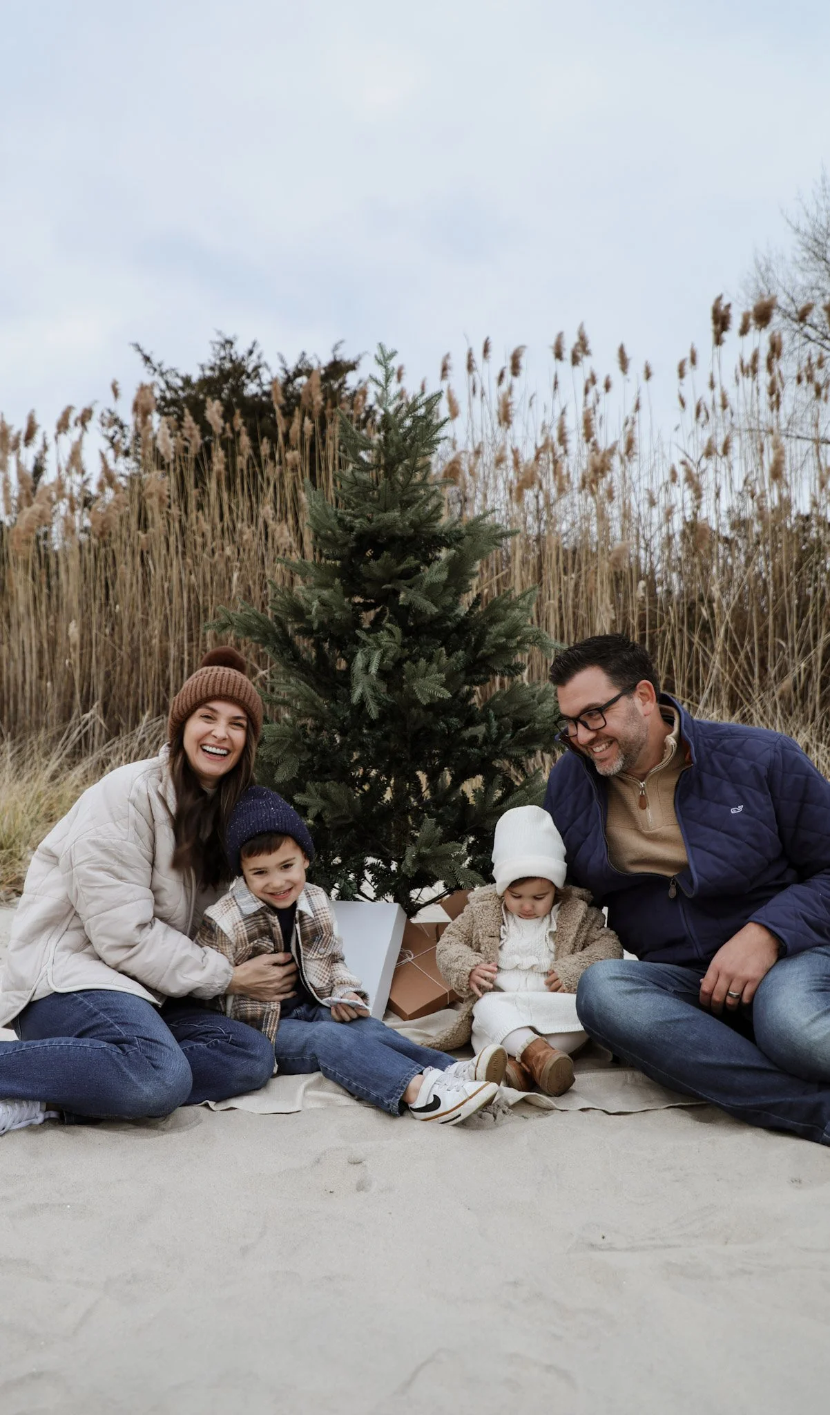 A family of four celebrating Christmas outdoors with a decorated Christmas tree, surrounded by tall dry grass and trees, on a cloudy day. The family is smiling and dressed warmly.
