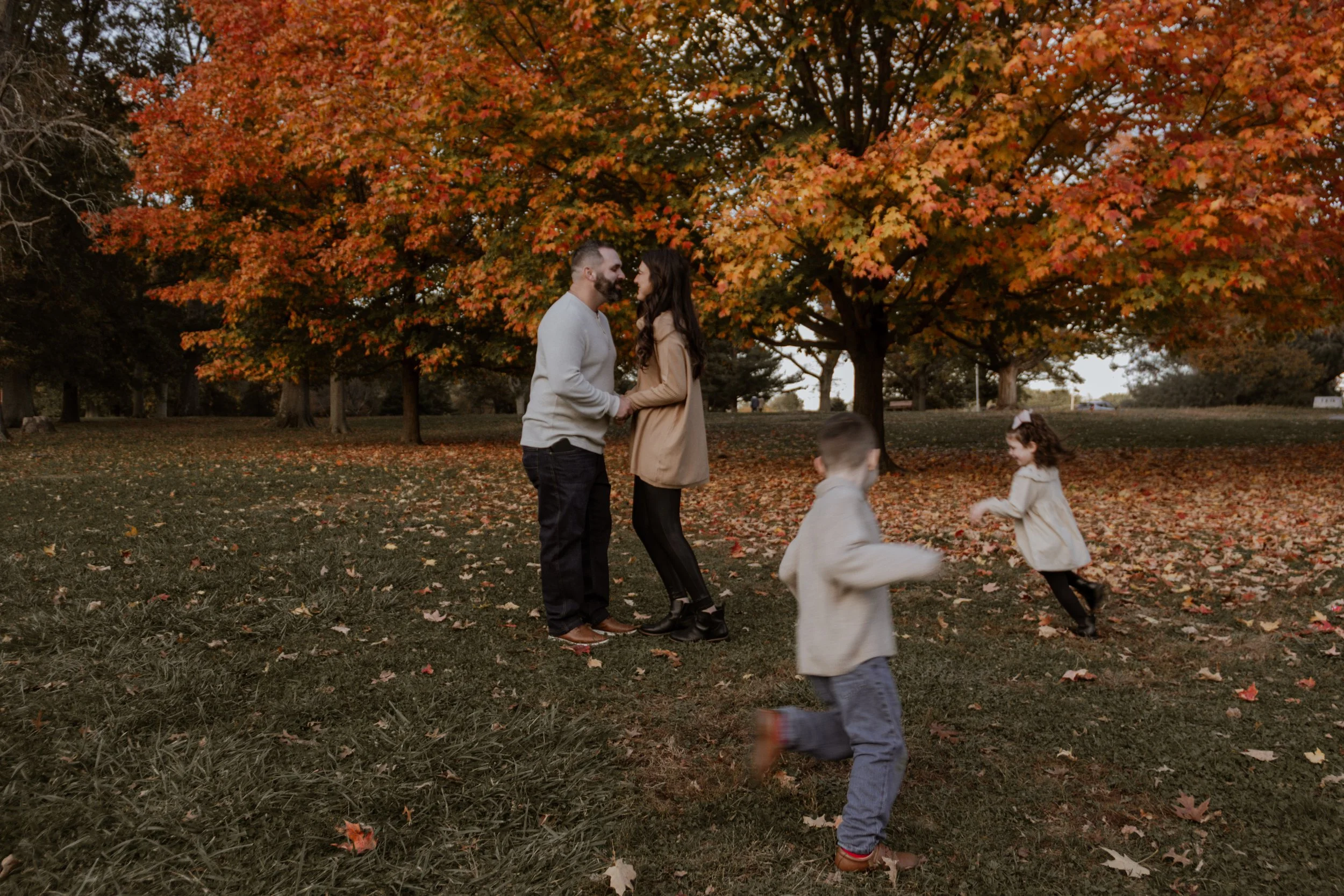 A couple holding hands and facing each other in a park with autumn trees and colorful leaves, while children run and play nearby.