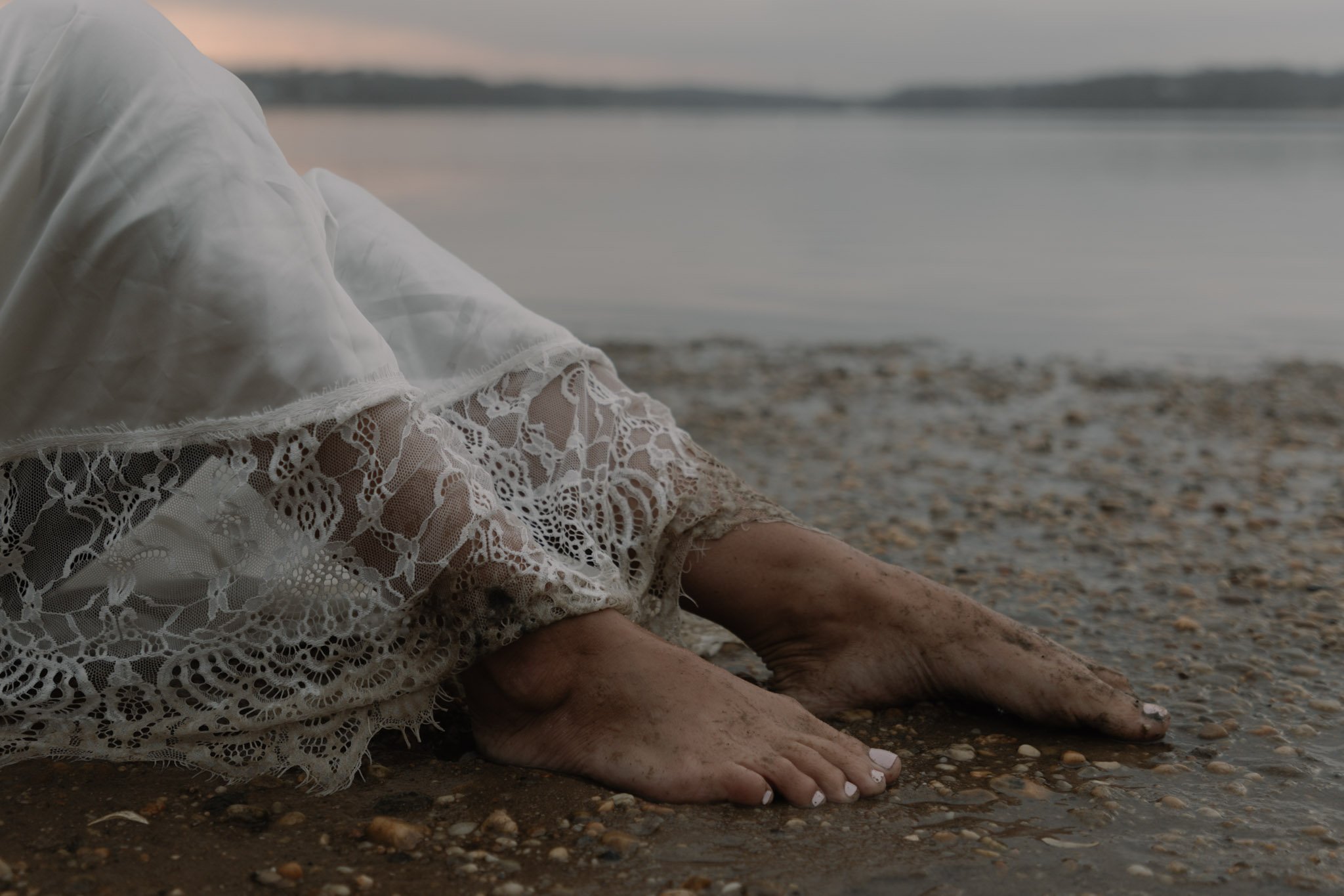 Close-up of a person's bare feet and lower legs resting on a pebbled beach, wearing a white lace dress, with a body of water and a cloudy sky in the background at sunset.