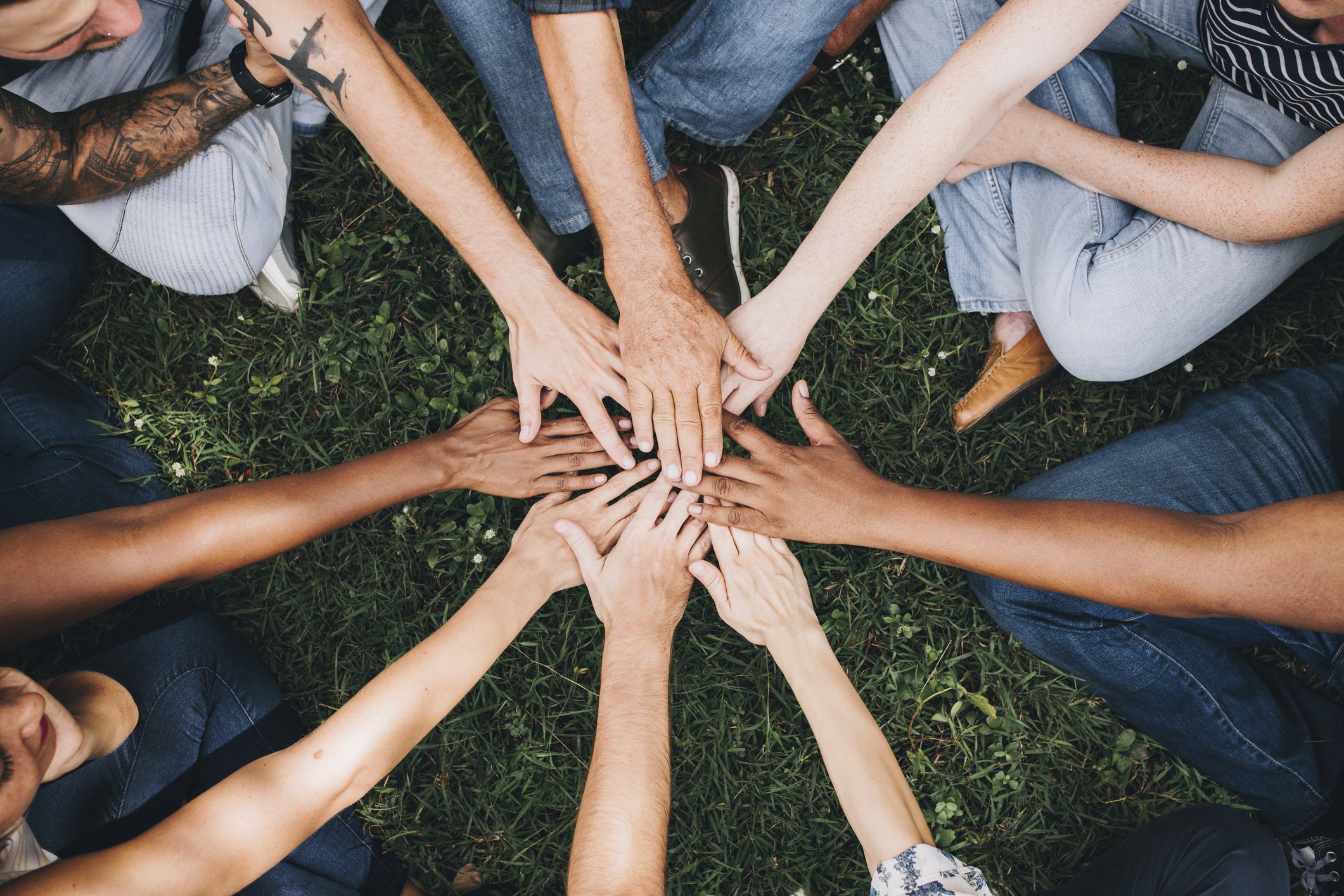 Multiple people sitting outdoors on grass, each extending a hand towards the center to create a joint hand stack, symbolizing teamwork or unity.