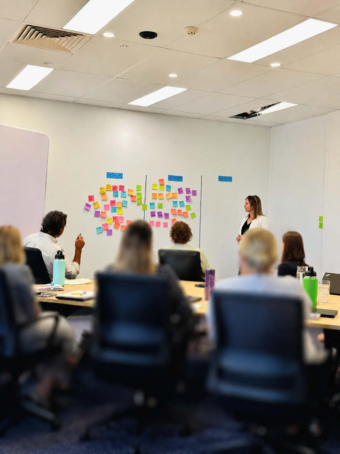 A woman is giving a presentation in a conference room with a whiteboard covered in colorful sticky notes. Several people are seated around a table listening attentively.