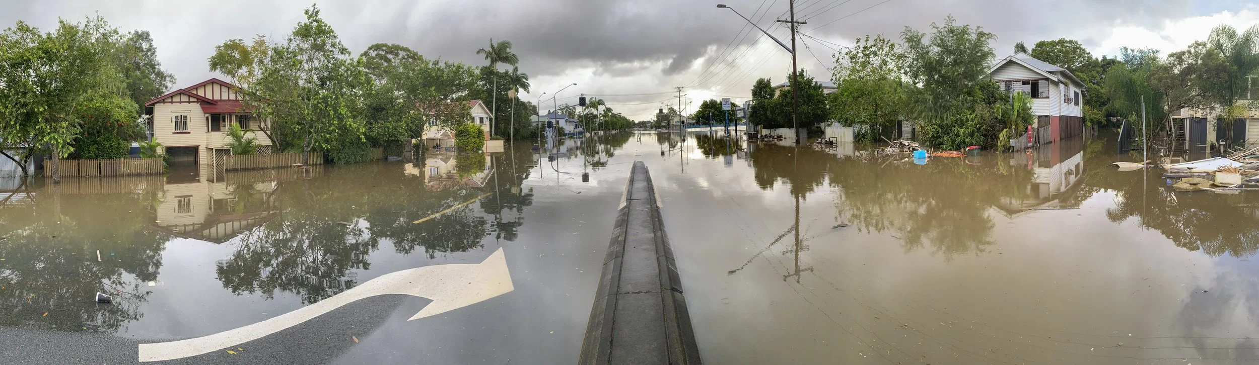 A flooded street with water covering the road and reaching the houses. Debris and personal items are floating in the water. Stormy weather with dark clouds overhead.