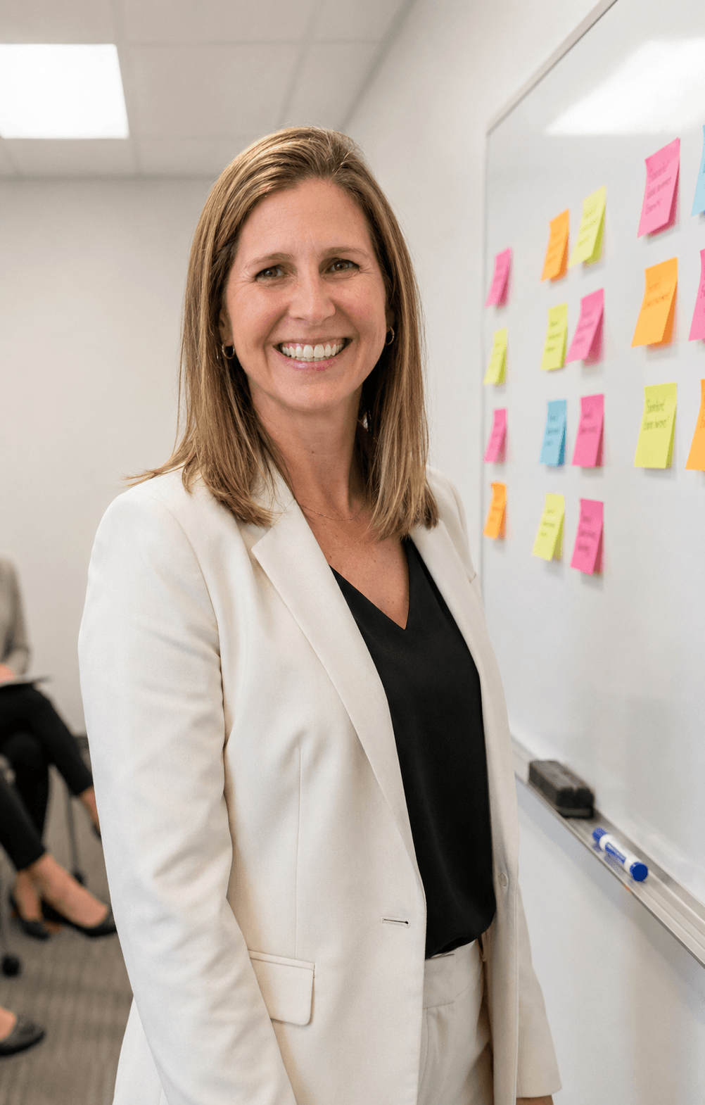 A woman with shoulder-length light brown hair, wearing a white blazer and black top, standing in front of a whiteboard with colorful sticky notes.