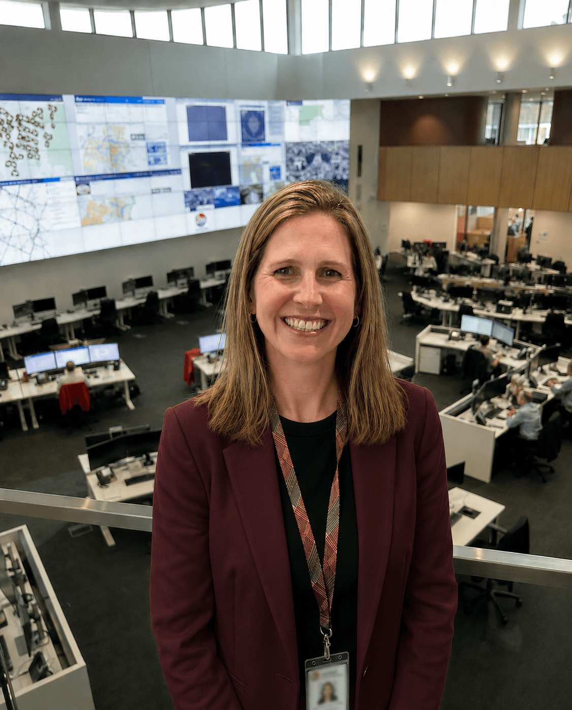 A woman smiling in a control room with multiple monitors and people working at computer desks.