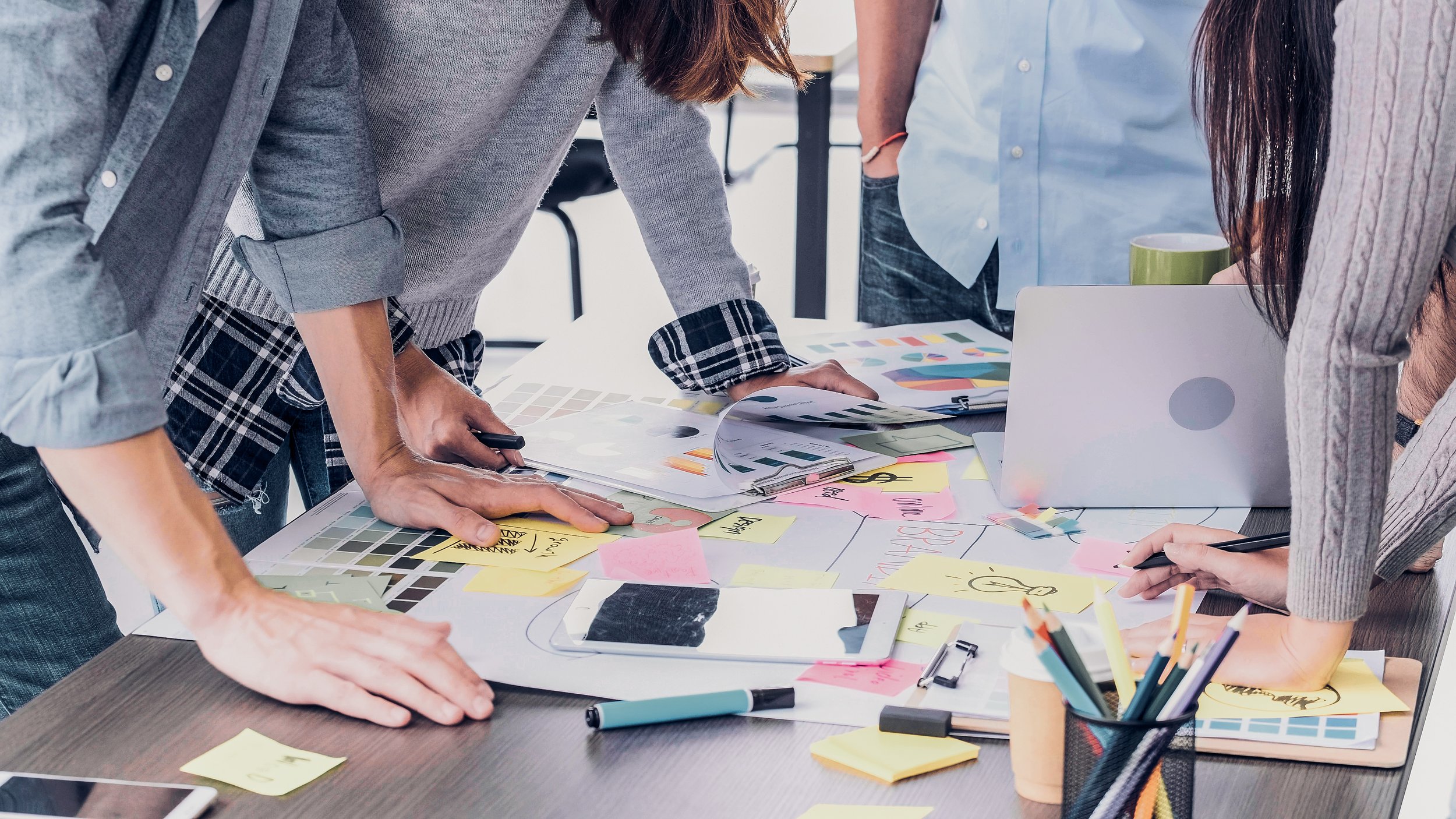 Group of people working together at a table covered with papers, sticky notes, and design materials, collaborating on a creative project.