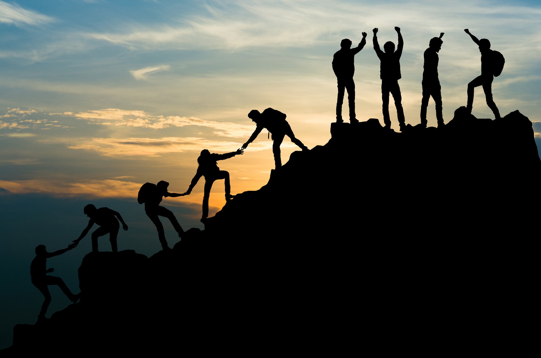 Silhouettes of people helping each other climb a mountain during sunset, with some reaching the top and others assisting each other.