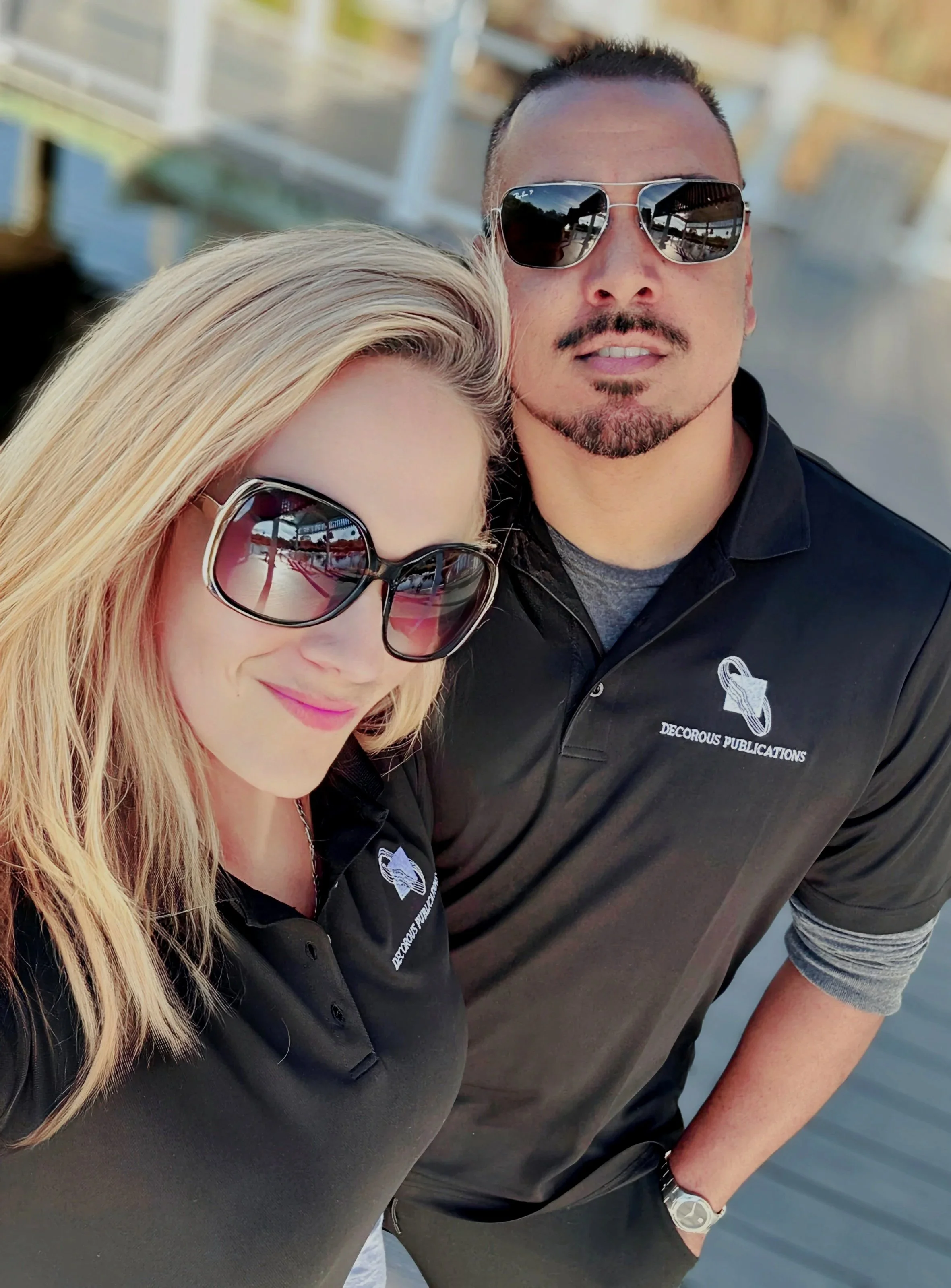 Close-up selfie of a man and woman, both wearing sunglasses, leaning towards each other on a bridge or outdoor walkway, with trees in the background.
