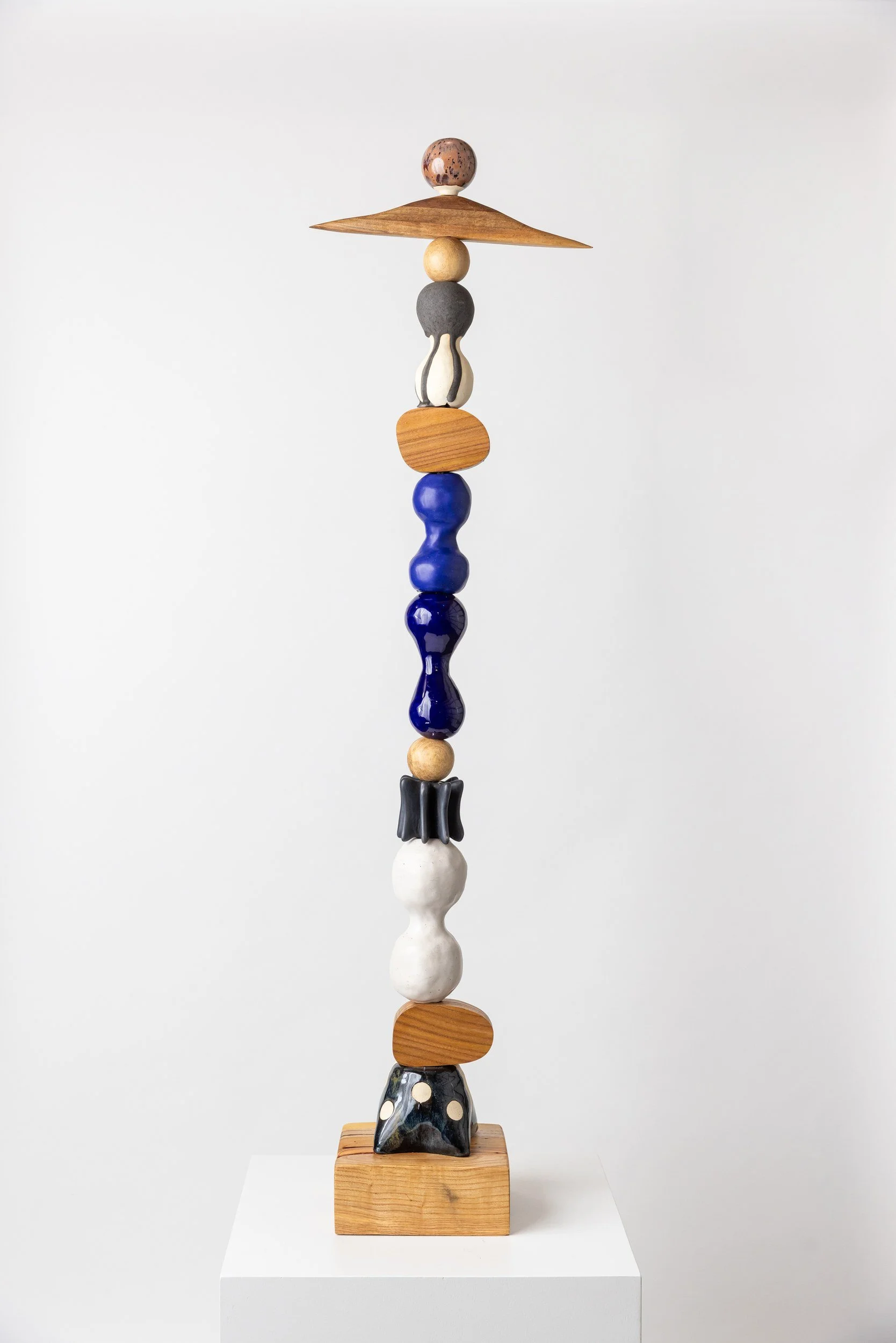 A tall sculpture made of various wooden and ceramic pieces stacked vertically on a wooden base against a plain white wall.