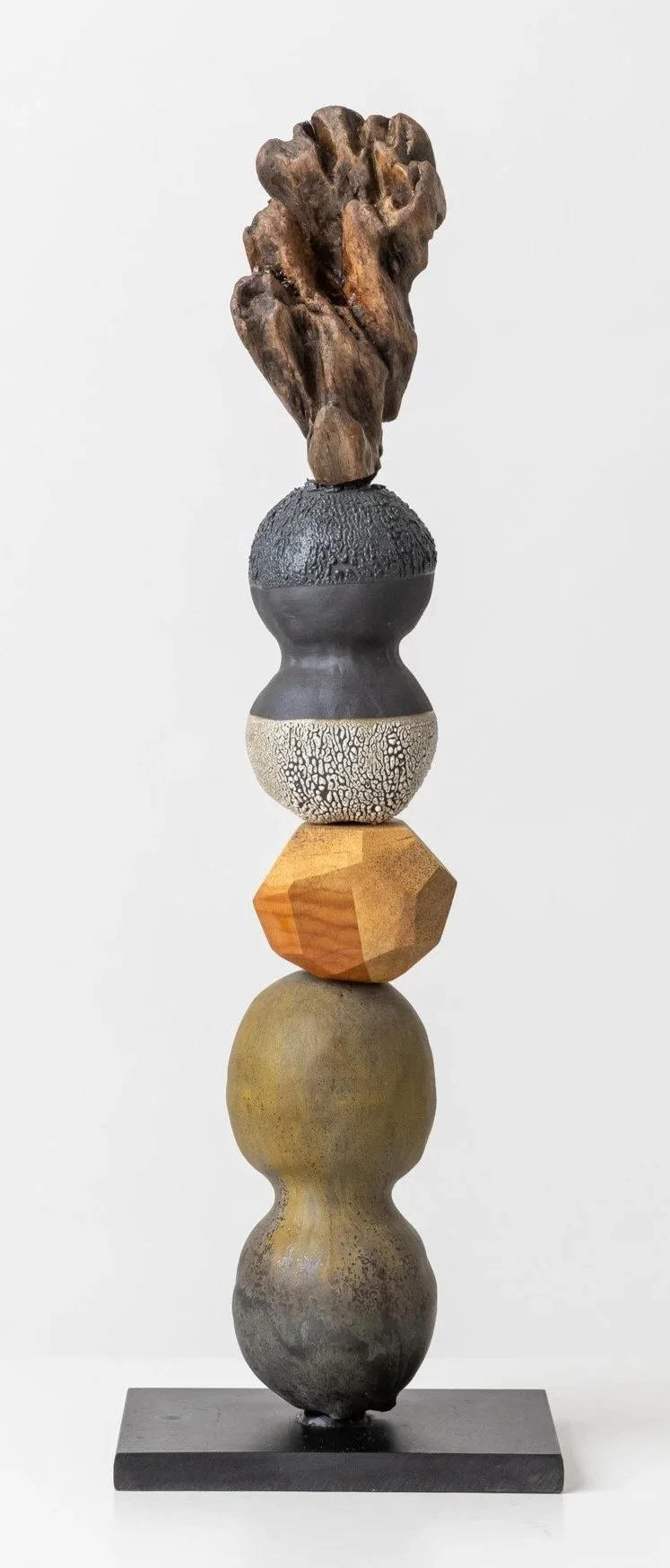 A tall sculpture made of stacked variously textured and colored stones, top to bottom: a rough dark brown piece, a gray textured sphere, a cream-colored sphere with a cracked pattern, a faceted wooden piece, and a rounded earthy-toned stone, mounted on a rectangular black base.