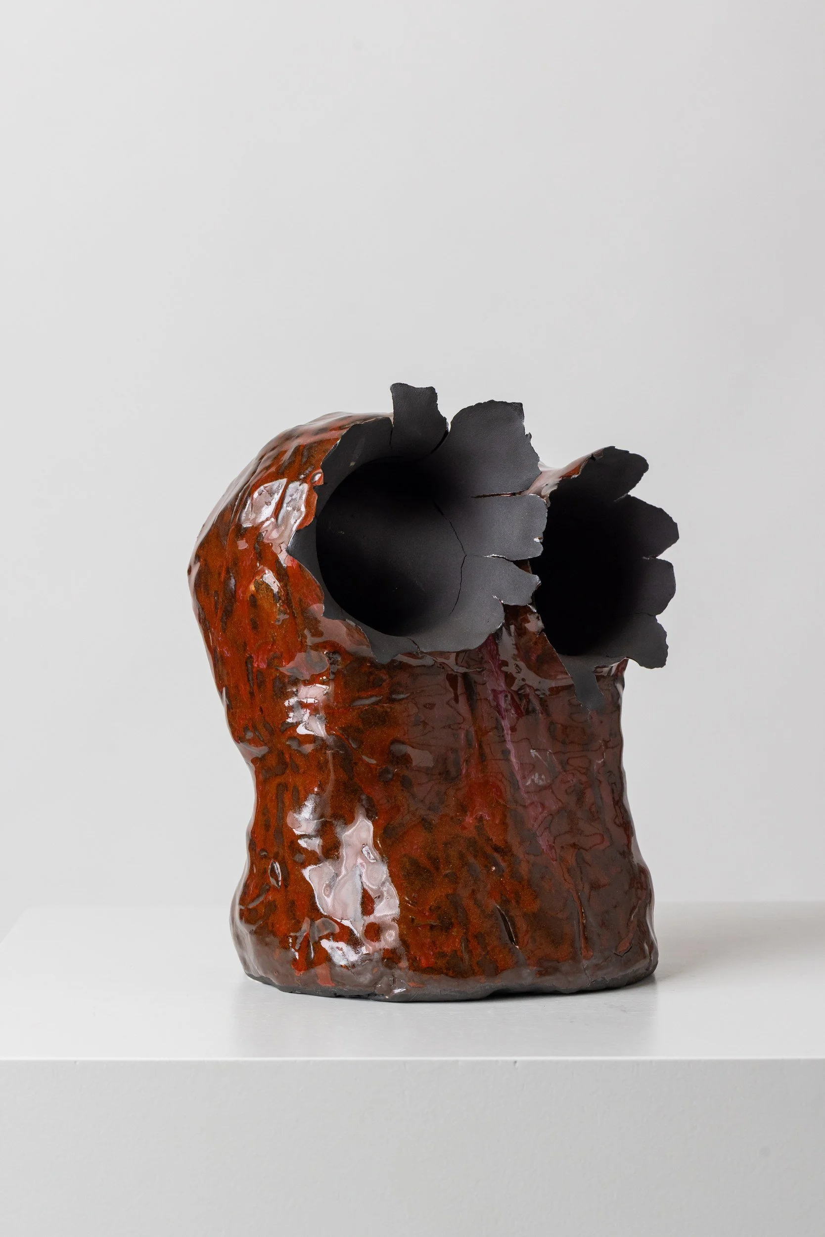 Ceramic sculpture resembling a flower with a reddish-brown textured exterior and dark interior petals, set on a white pedestal against a plain white background.