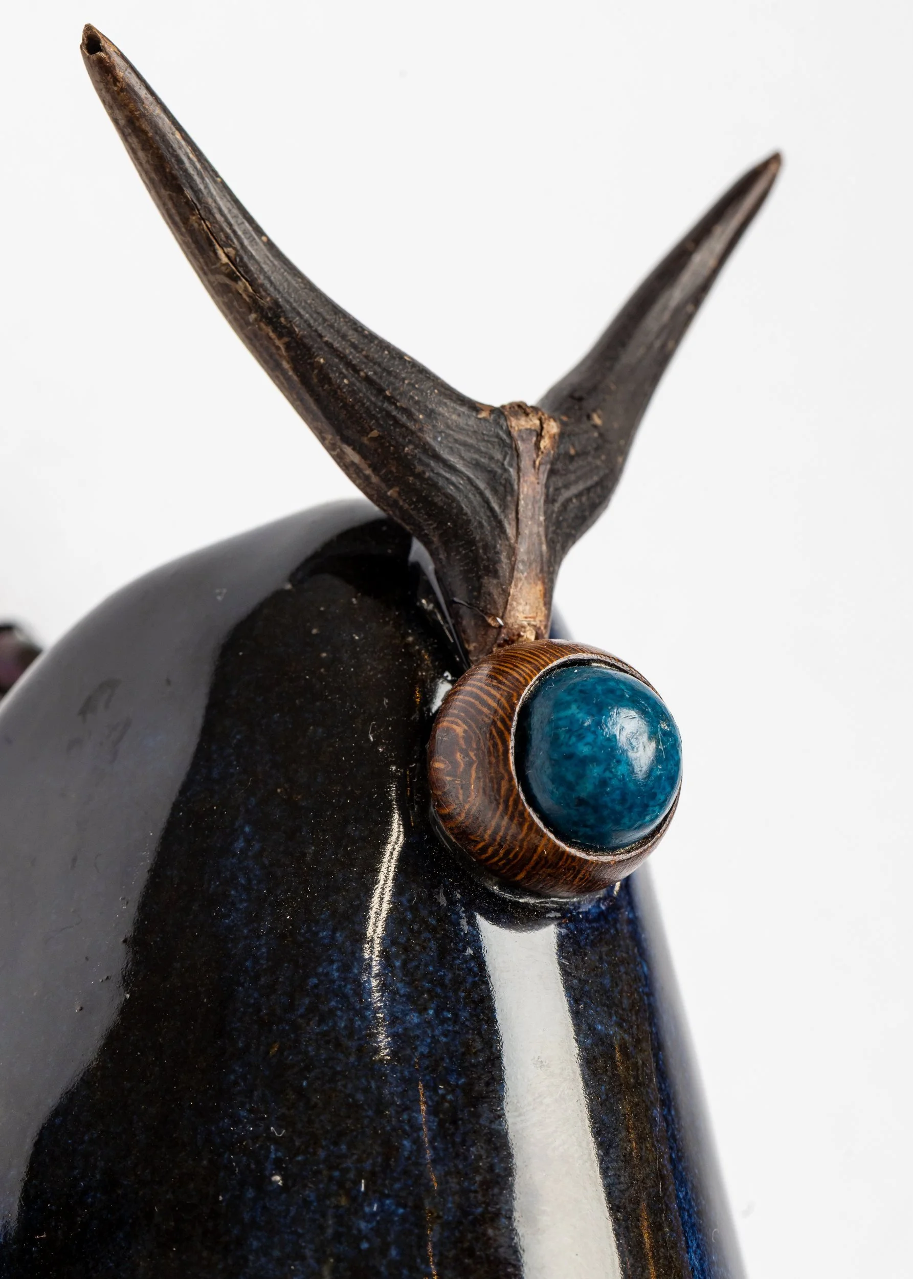 Close-up of a dark-colored sculpture with a wooden element and a blue stone, against a white background.
