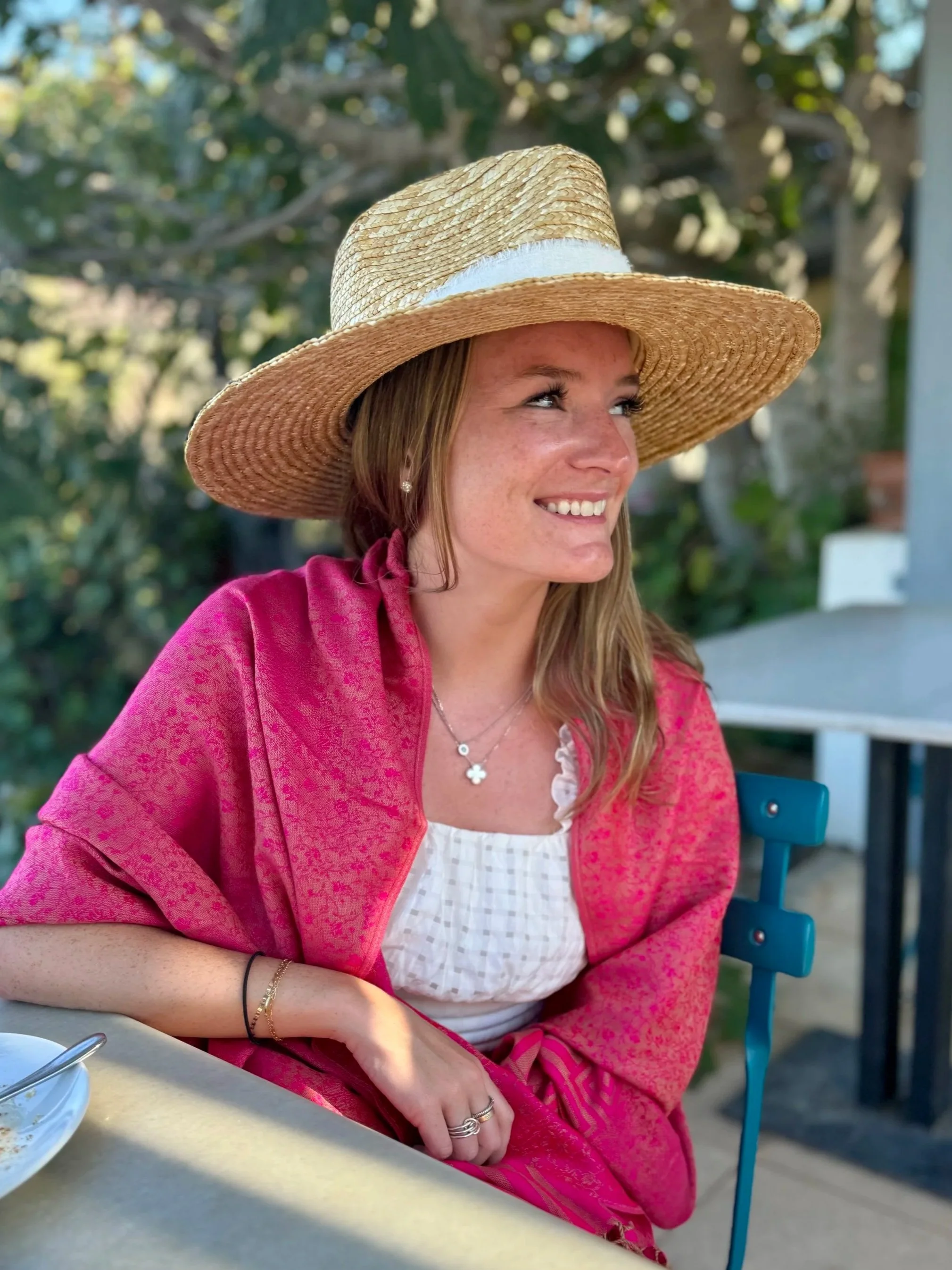 A woman wearing a large straw sunhat, pink shawl, and white top sitting at an outdoor table with a smile, surrounded by greenery.