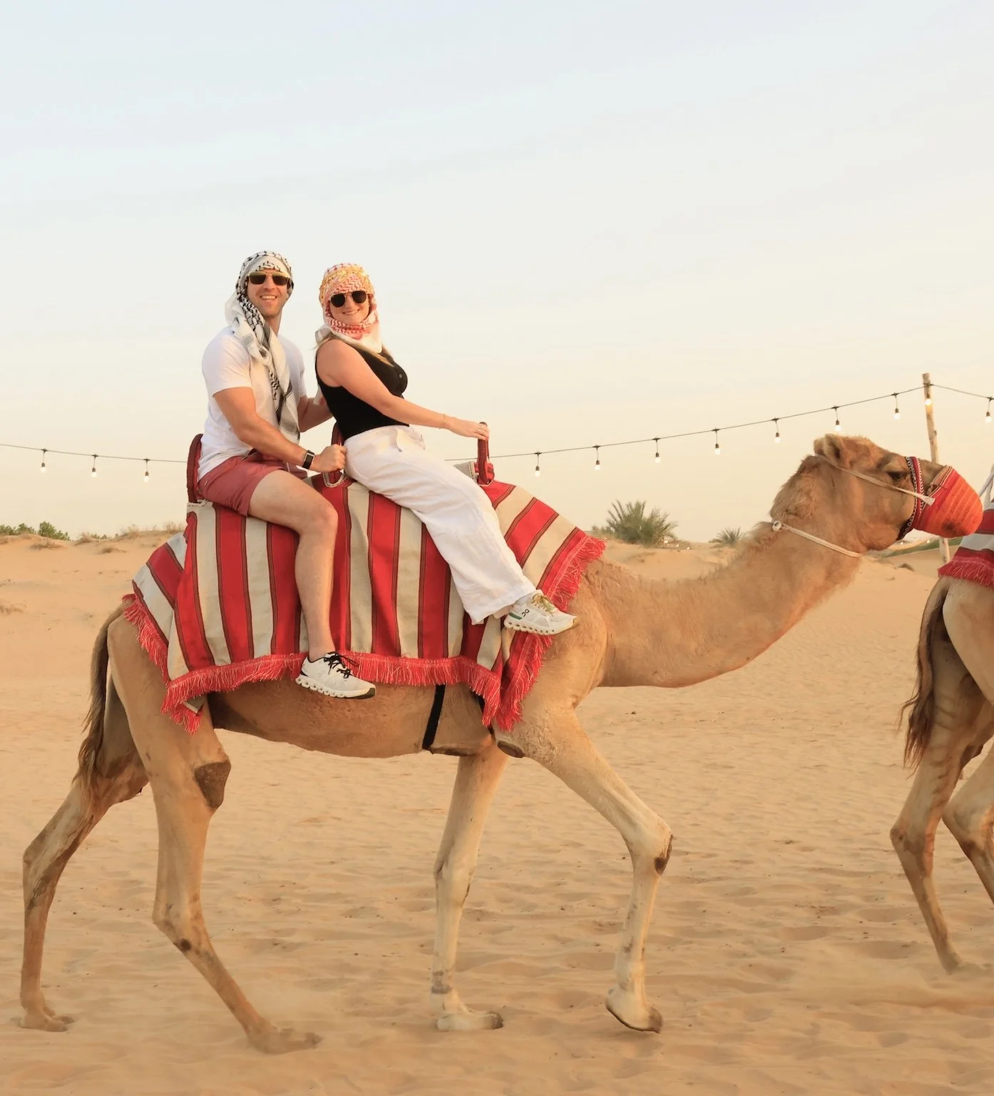 Two people riding a camel in a desert at sunset, wearing sunglasses and headscarves, with string lights in the background.