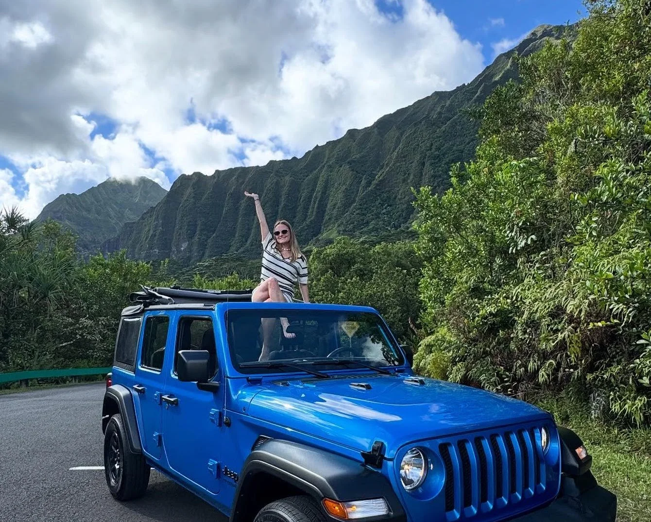 A woman sitting on top of a blue Jeep Wrangler on a mountain road, with lush green mountains and partly cloudy sky in the background.