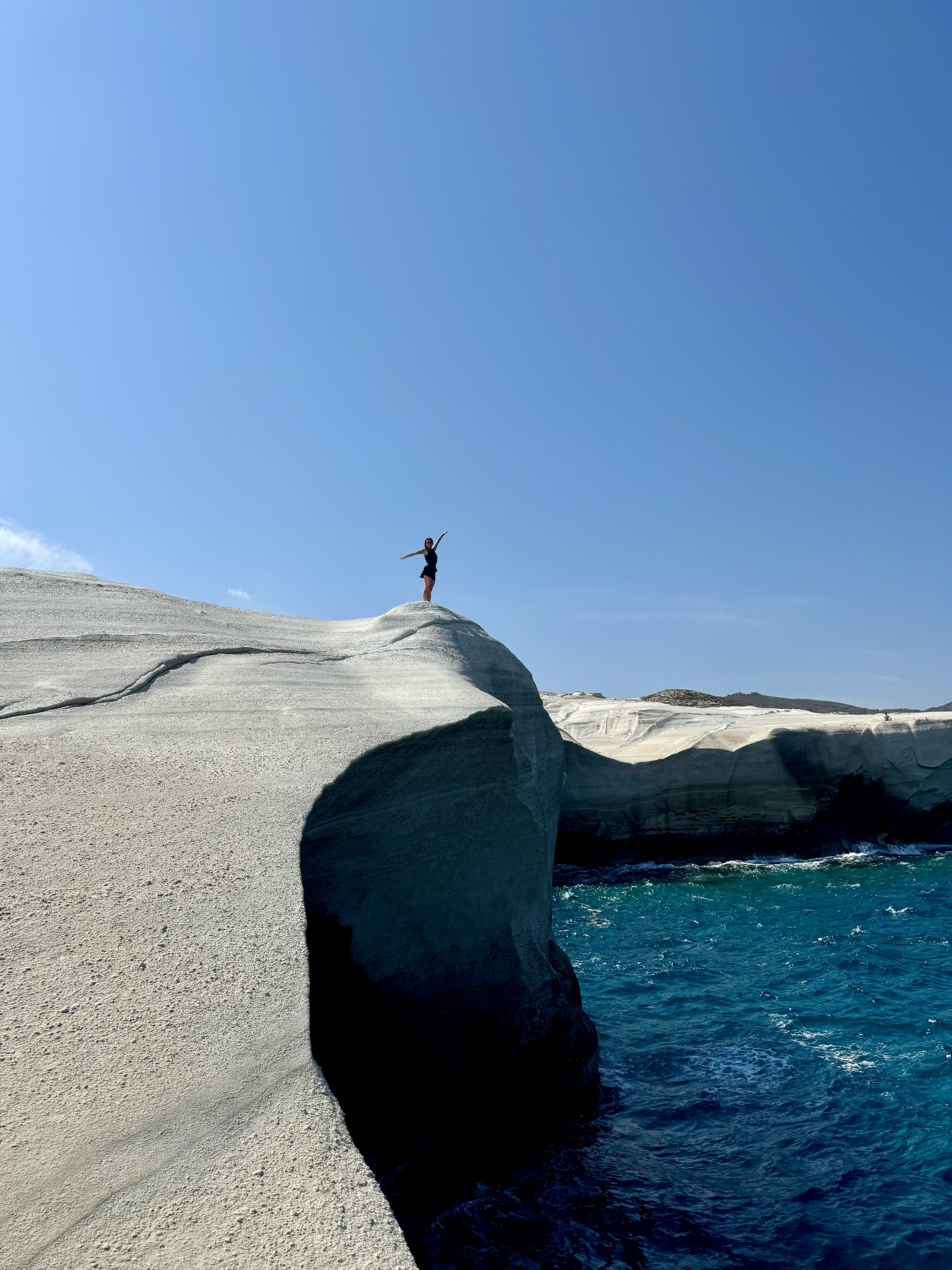 A person standing on a white rock formation by the sea, with one arm raised in a swirl, under a clear blue sky.