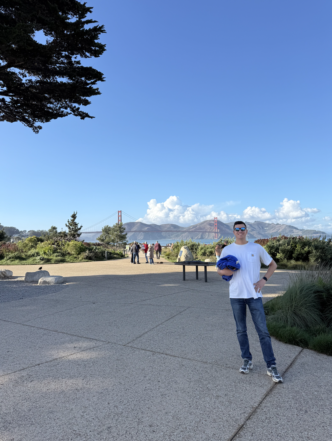 A young man wearing sunglasses, a white t-shirt, and jeans stands on a paved area near a park, holding a blue jacket. The Golden Gate Bridge is visible in the background with the hills behind it, and several people are walking and standing around the park on a clear day.