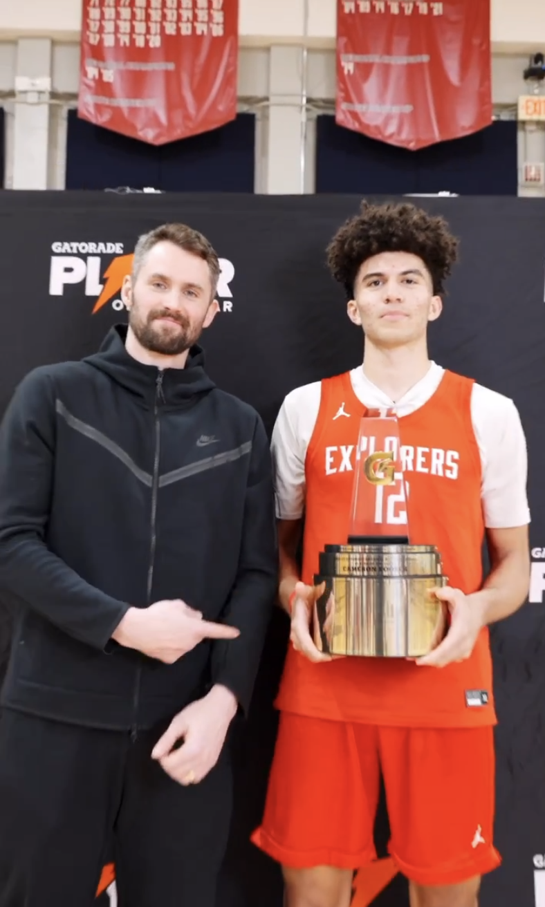 A young basketball player in a red jersey holding a trophy, standing next to a man pointing at him, indoors at a basketball event.