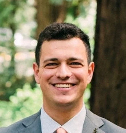 A smiling man in a suit outdoors with trees in the background.