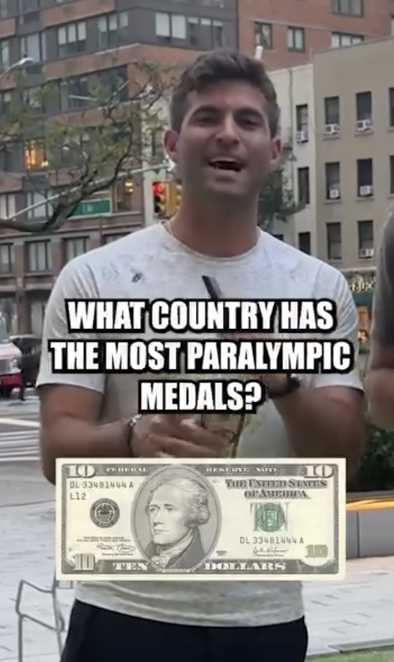 A young man standing on a city street holding a pen and a paper, with a dollar bill superimposed in front of him. The text overlay asks, "What country has the most Paralympic medals?"