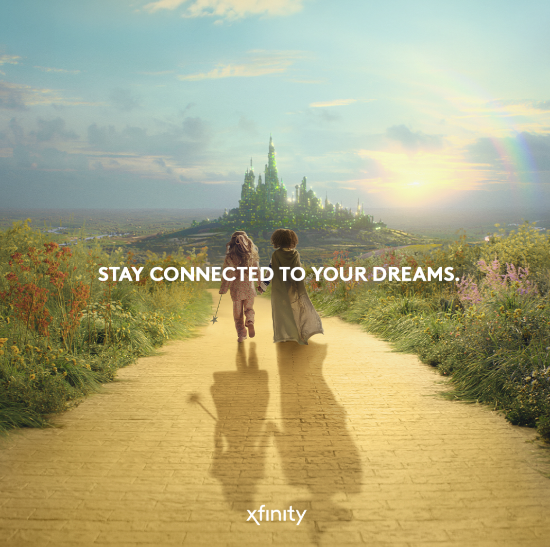 Two children, dressed as a princess and a wizard, walk hand in hand down a yellow brick road surrounded by colorful flowers towards a castle in the distance under a bright sky. The text reads "Stay connected to your dreams."