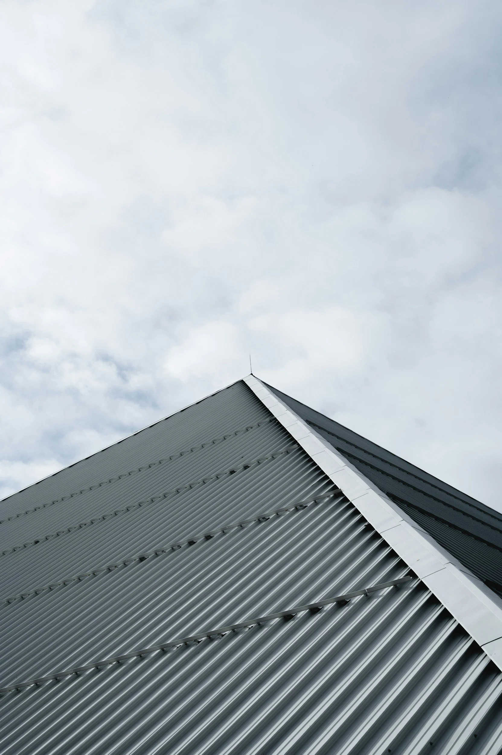 Close-up view of a metal roof with ridges and a sharp angular edge against a cloudy sky.