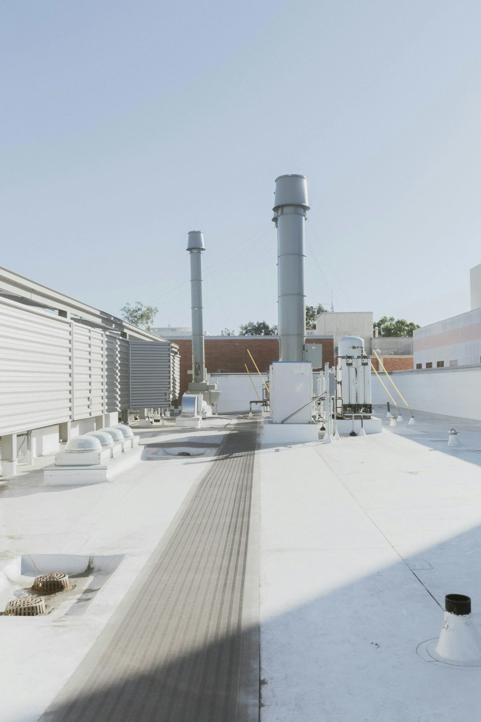 Rooftop with white concrete surface, ventilation pipes, electrical equipment, and a grey walking path with drainage grates under a clear blue sky.