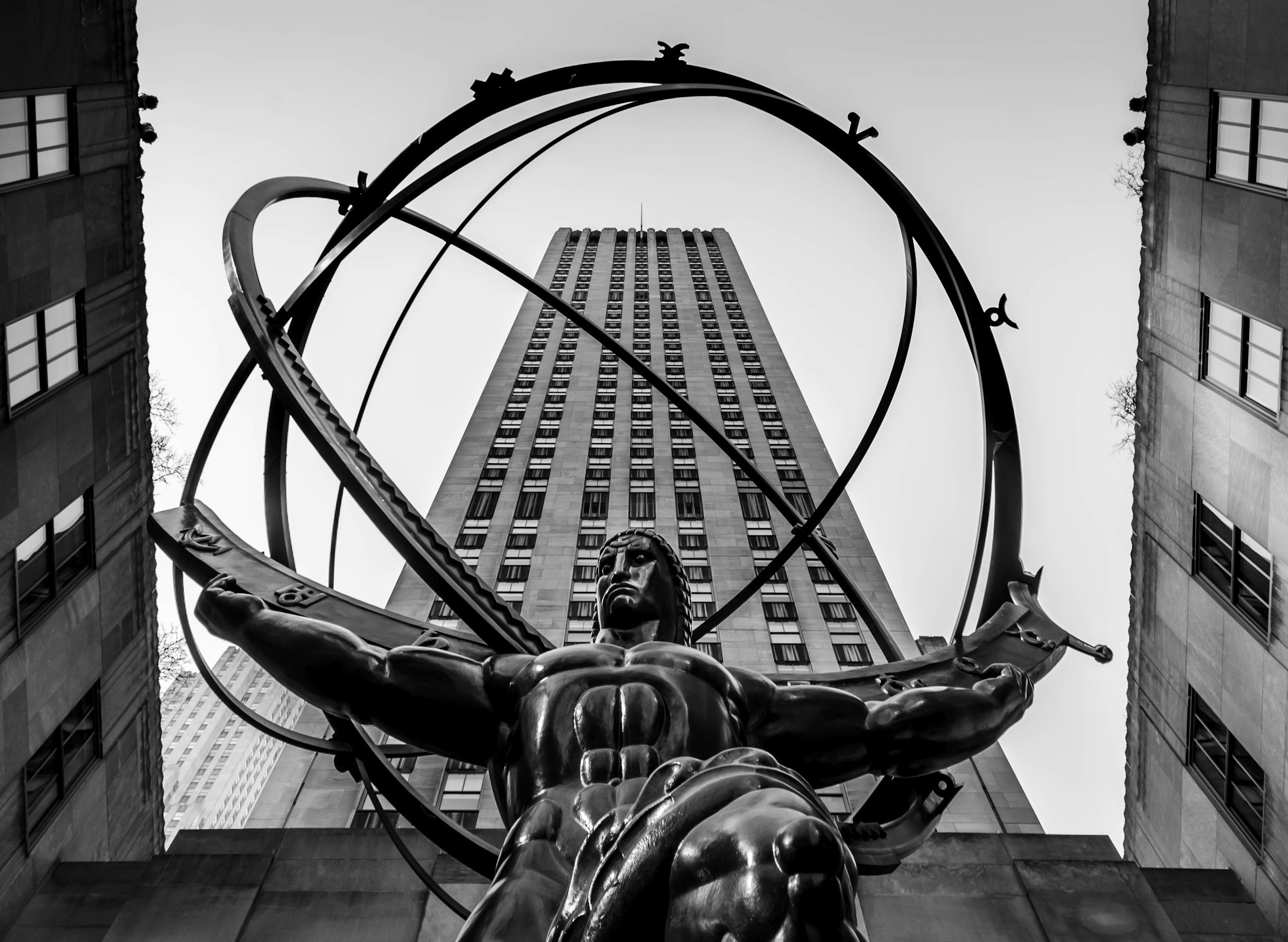Black and white photograph of a tall building viewed from the ground, with a large metallic sculpture in the foreground featuring a human figure holding an armillary sphere.