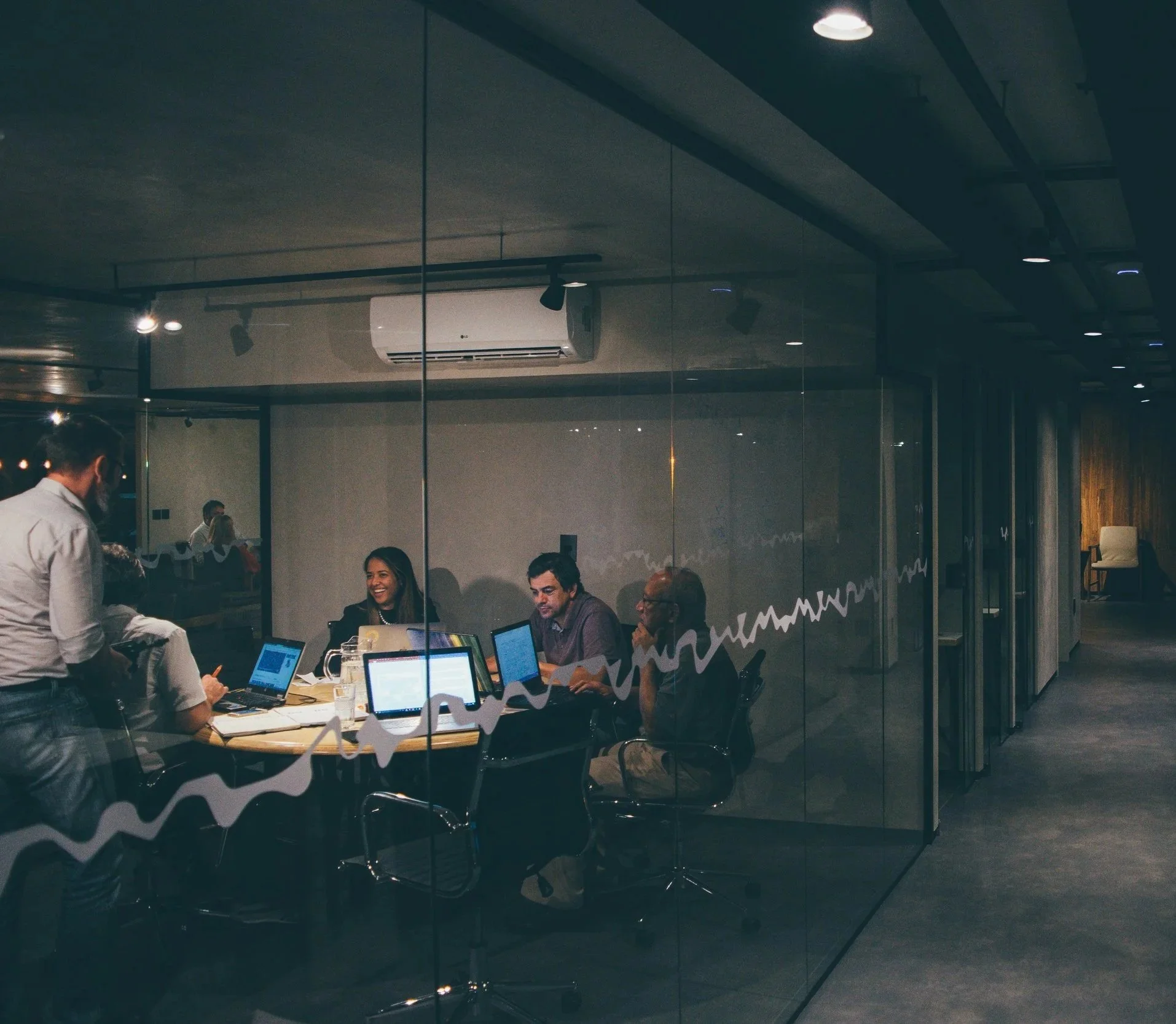 A business meeting or workshop with people working on laptops at a round table, seen through a glass wall with decorative patterns, in a modern conference room.