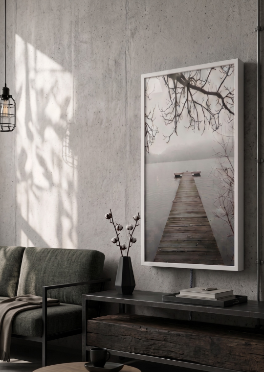 Interior of a modern room with a large framed photograph of a dock extending into a calm lake, bare tree branches overhead, a green sofa, a black side table with a vase and books, and a pendant light casting shadows on a concrete wall.