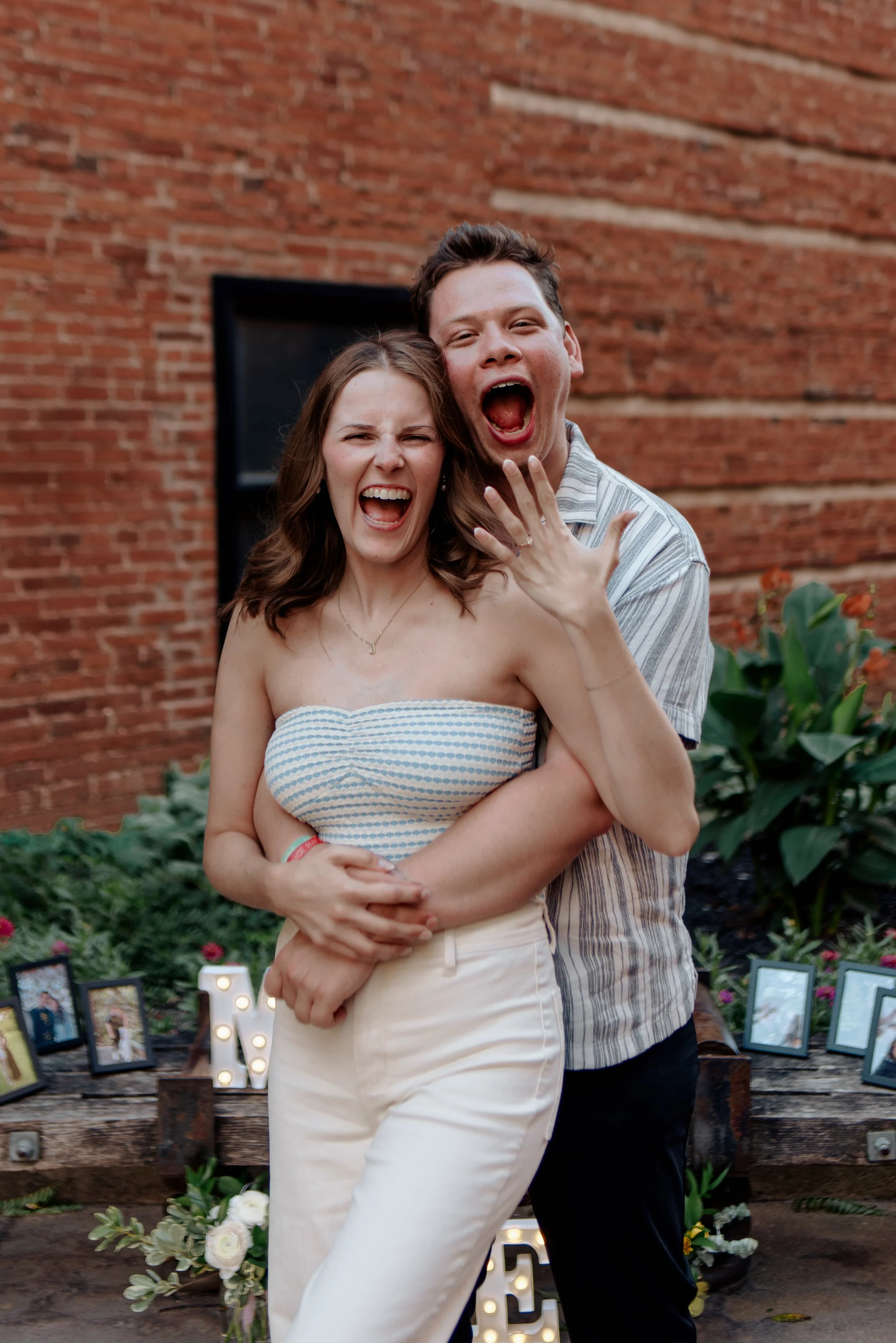 A joyful couple embraces in an outdoor setting with a brick wall background, decorated with framed photos, lit letters, and flowers.