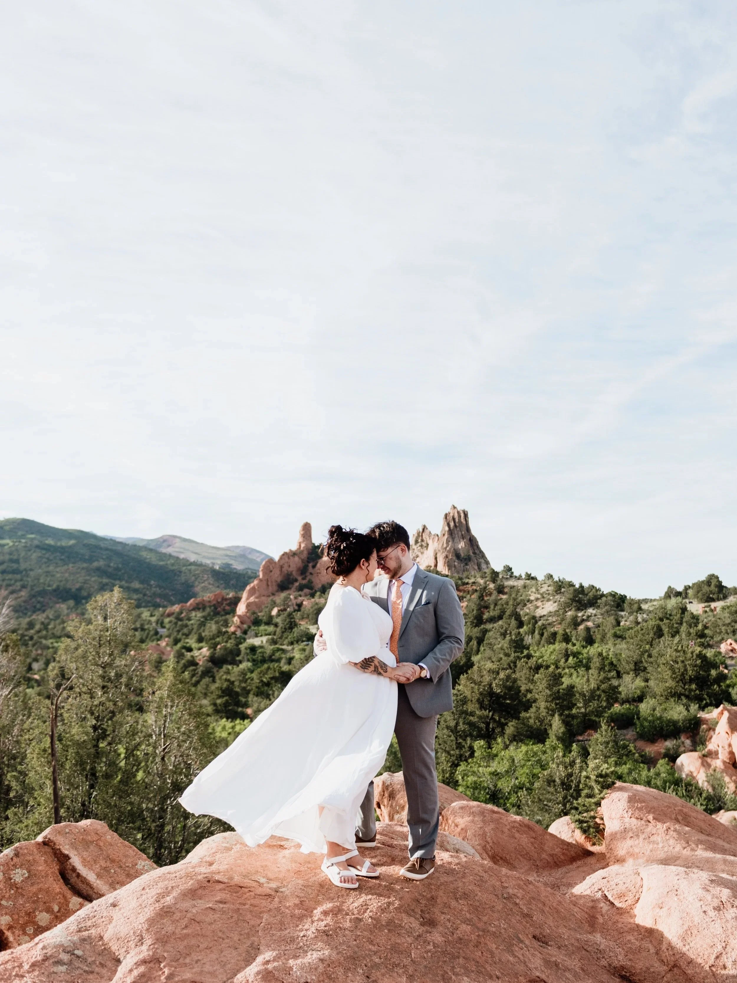A couple in wedding attire standing close together on a rocky outcrop with a scenic mountainous landscape and rock formations in the background.