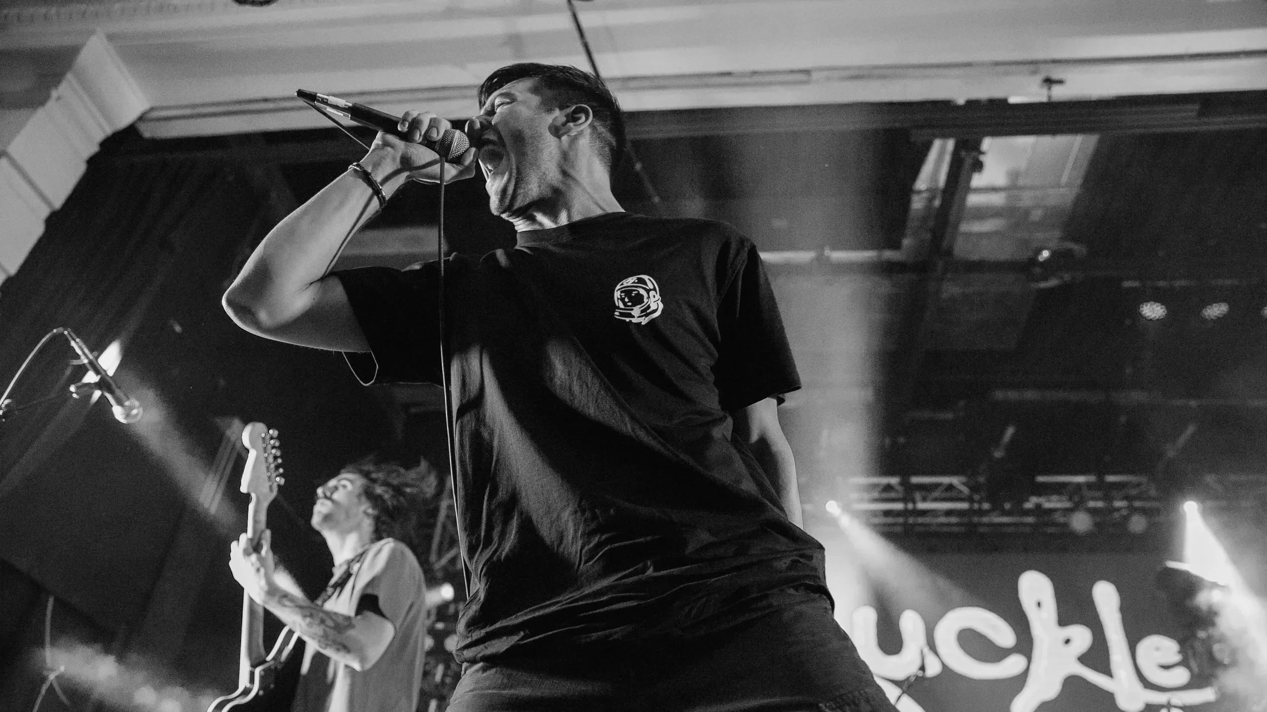 A black and white photo of Knucklepuck passionately singing into a microphone on stage, with a guitarist in the background.