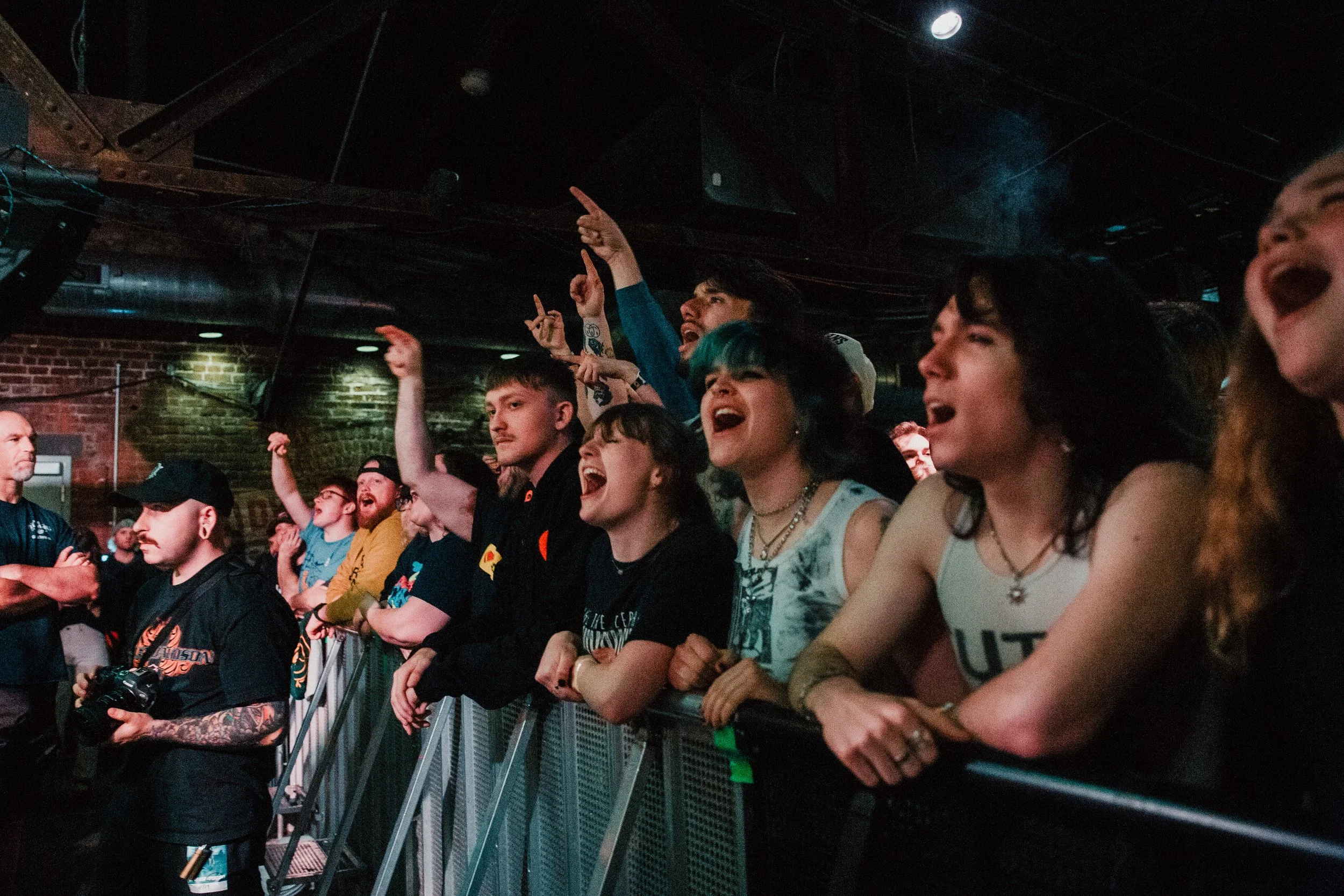 A crowd of young people at a concert, some singing, cheering, and pointing towards the stage, with a brick wall and metal barriers in the background.