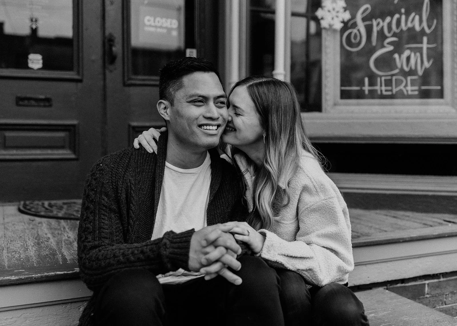 A young couple sitting on a bench outside a storefront. The woman is whispering into the man's ear, both smiling. 
