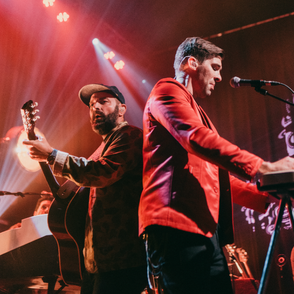 Drew Holcomb & the Neighbors performing on stage, one playing an acoustic guitar and the other using a keyboard, under stage lights.