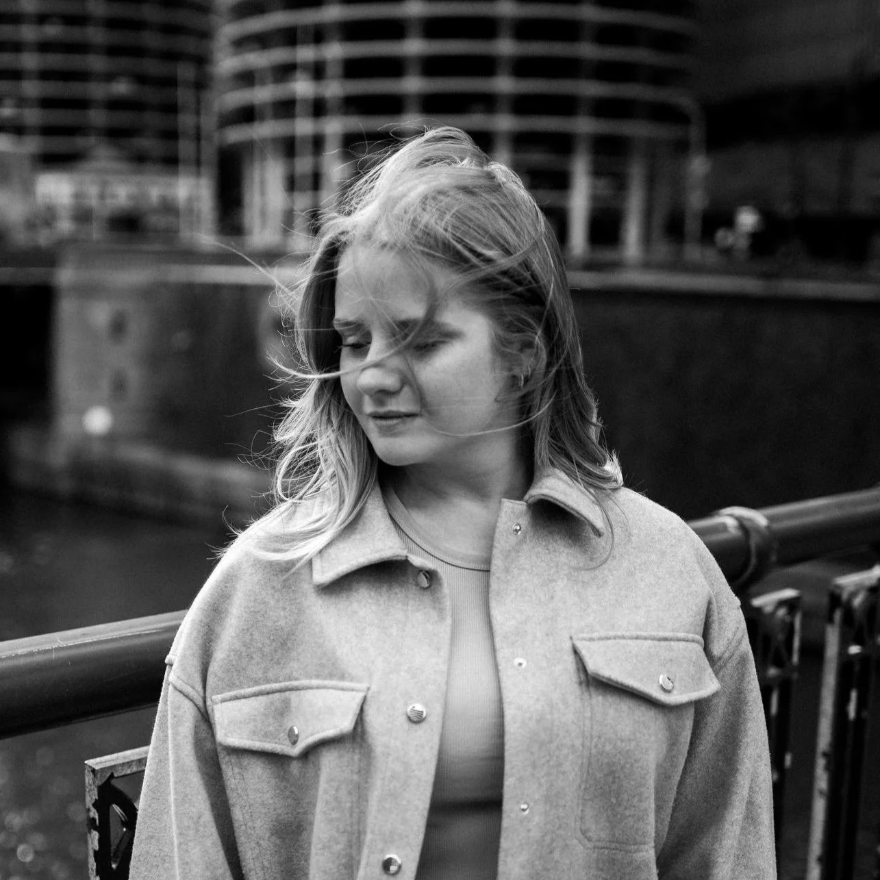 A black-and-white photo of a young woman with light hair, eyes closed, wind blowing her hair, standing outdoors near a railing with modern buildings in the background.