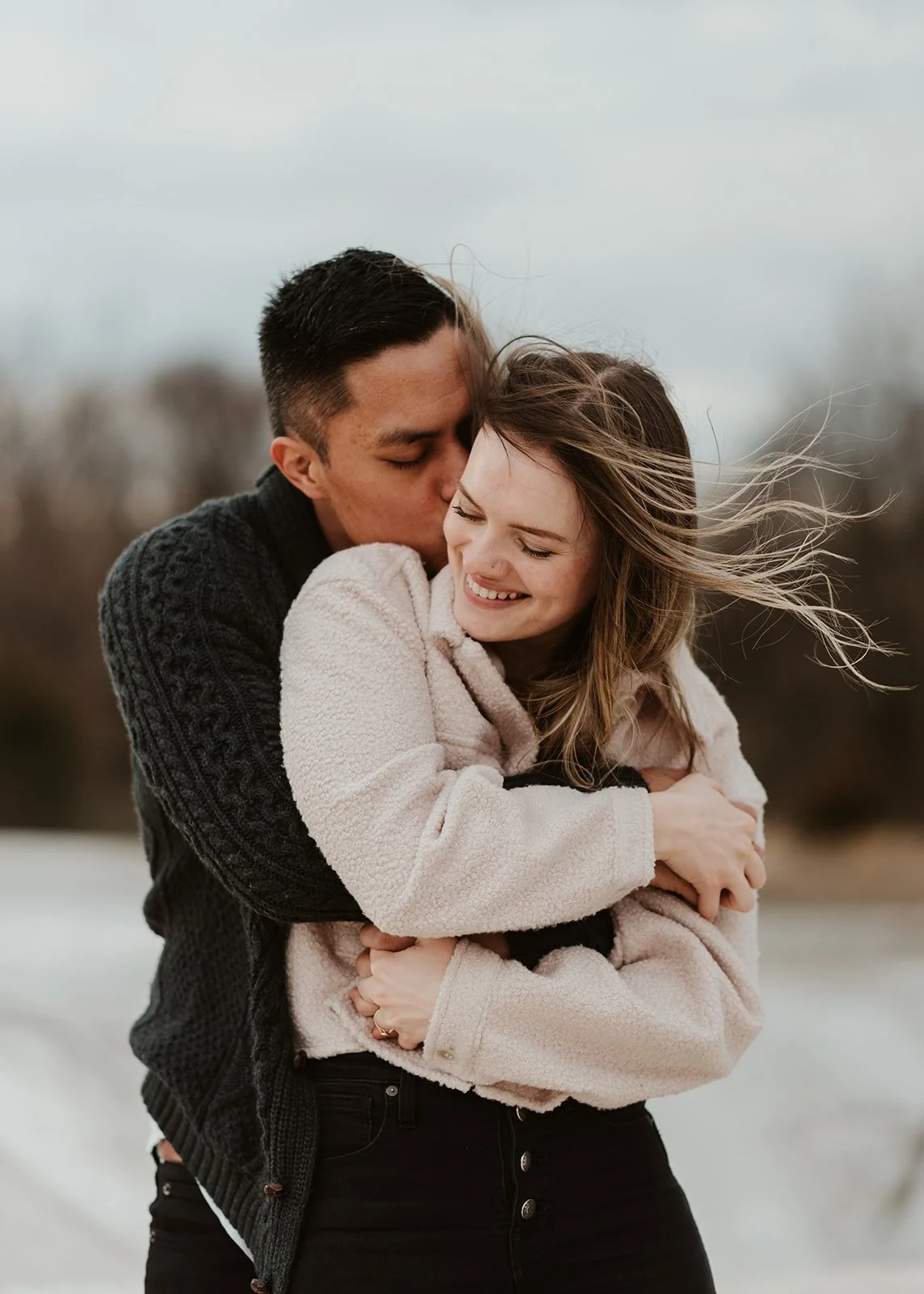A couple hugging outdoors on a windy day, the man kissing the woman on her forehead and the woman smiling with her eyes closed. Wind-blown hair and a cloudy sky in the background.