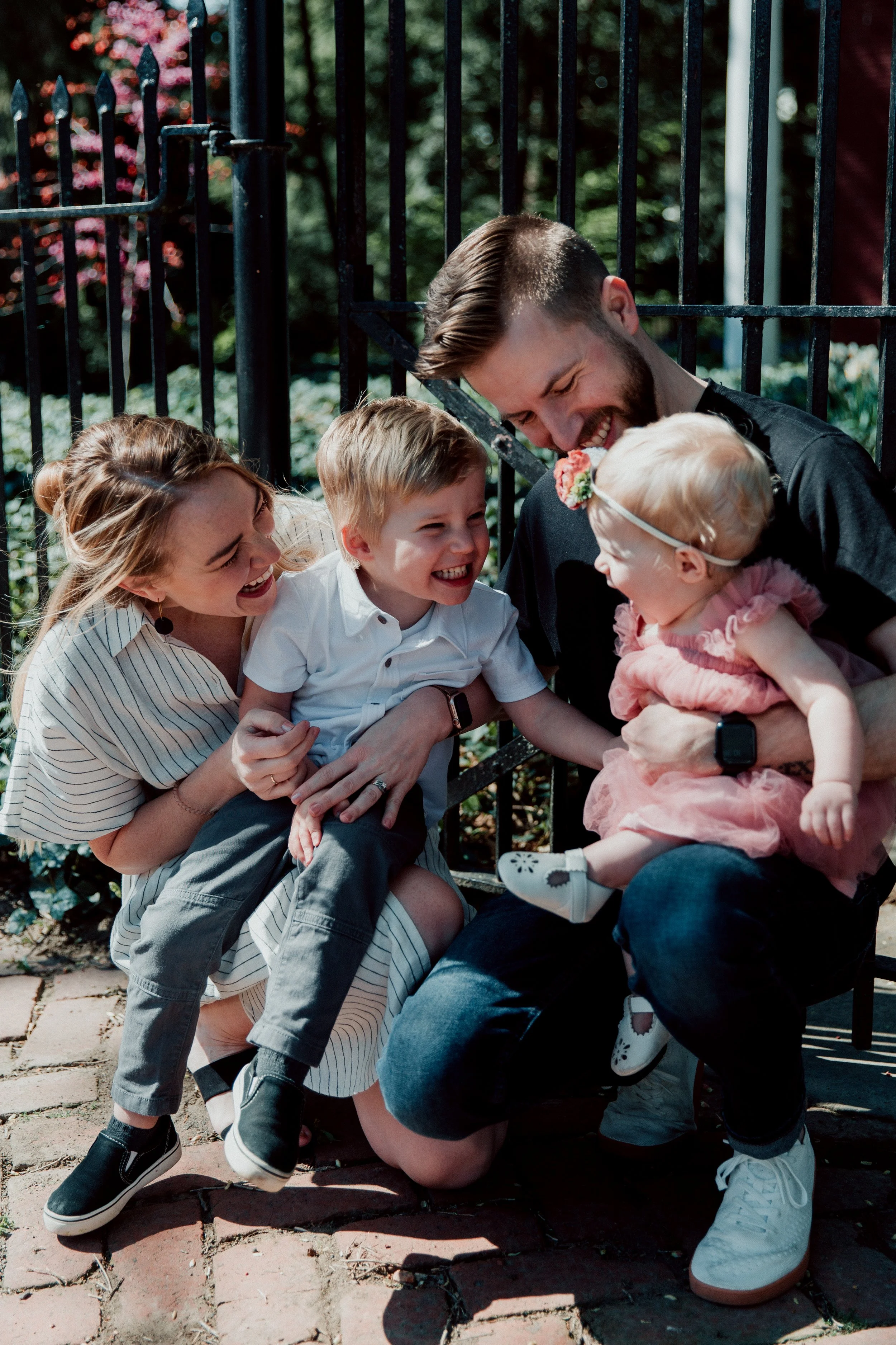 A family of four joyfully laughing and sharing a moment outside near a black metal fence, with greenery and trees in the background.